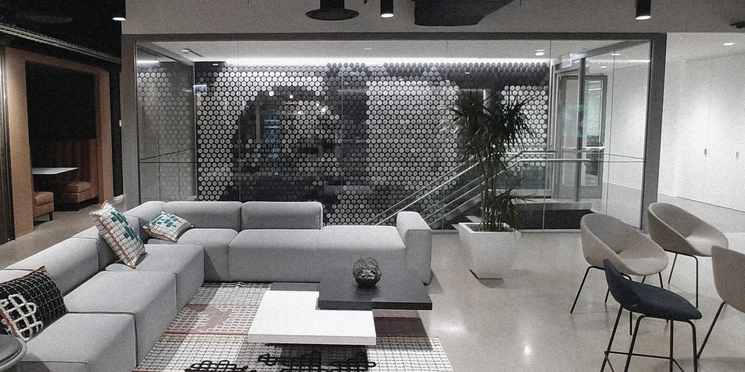 Modern lobby area with a large light gray sectional sofa decorated with patterned throw pillows, a black coffee table, and a white side table on a patterned rug. There are several chairs, a tall plant in a white pot, and a glass wall with a perforate