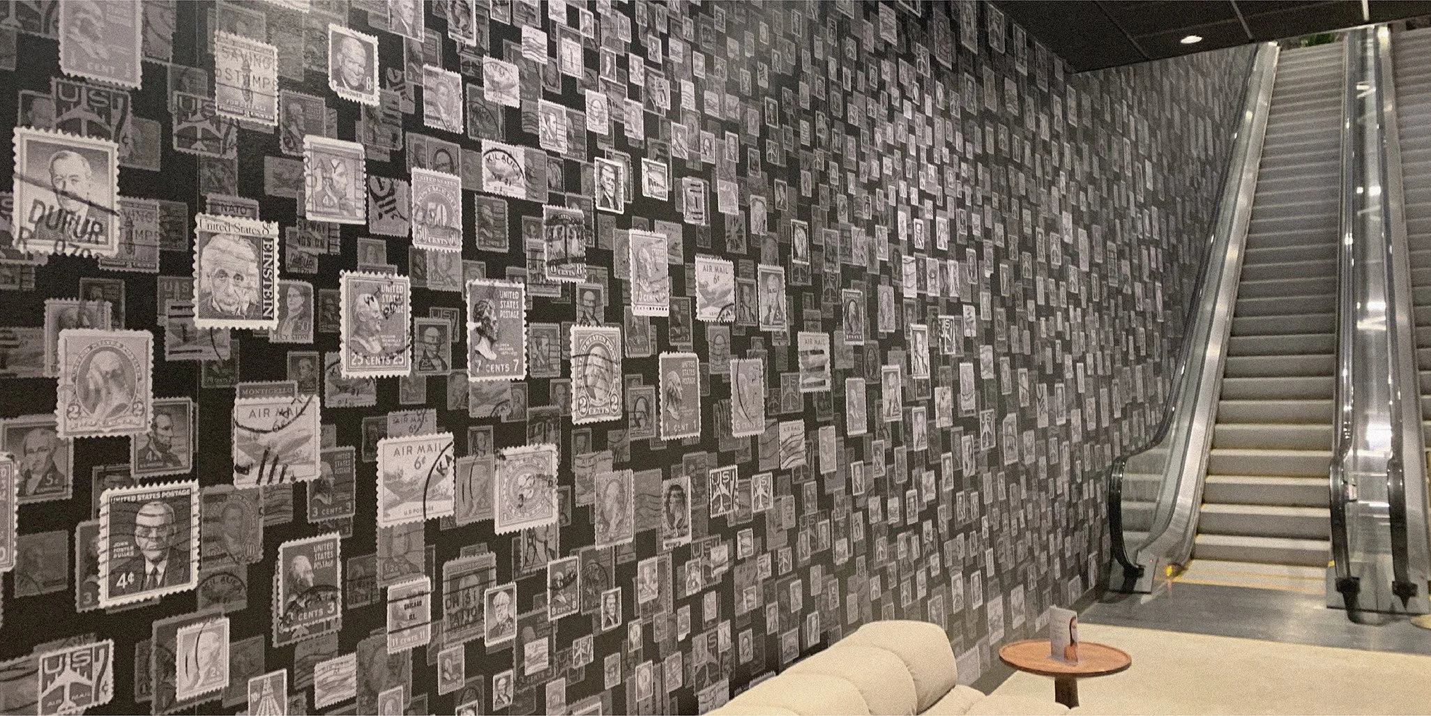 Interior of a building with a black wall covered in vintage postage stamp artwork, an escalator, a beige sofa, and a small round wooden table with a promotional card on it.