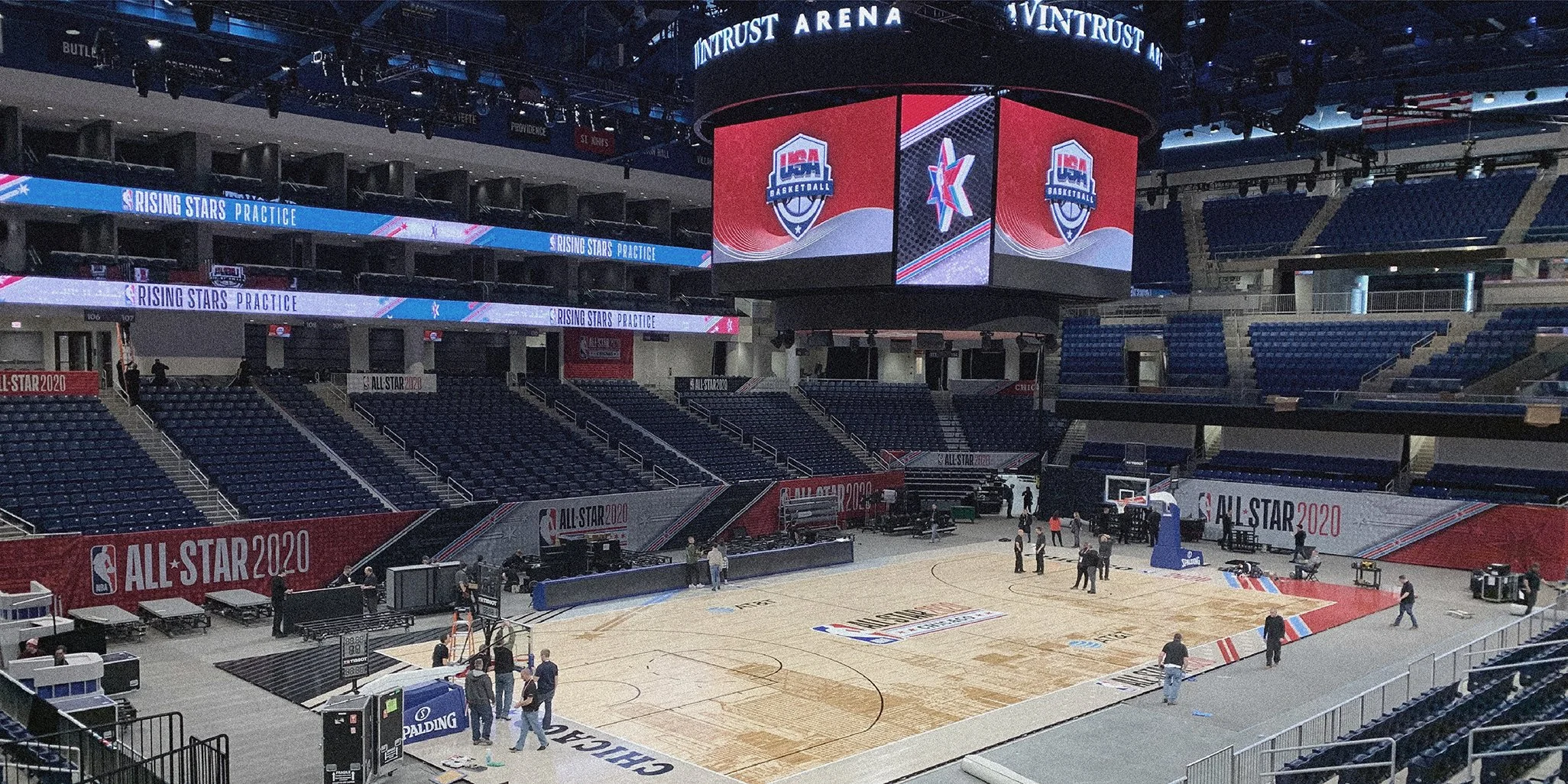 Empty basketball court inside a stadium with NBA All-Star 2020 branding, large digital screens displaying NBA Rising Stars Practice, and team logos, with some people setting up equipment.