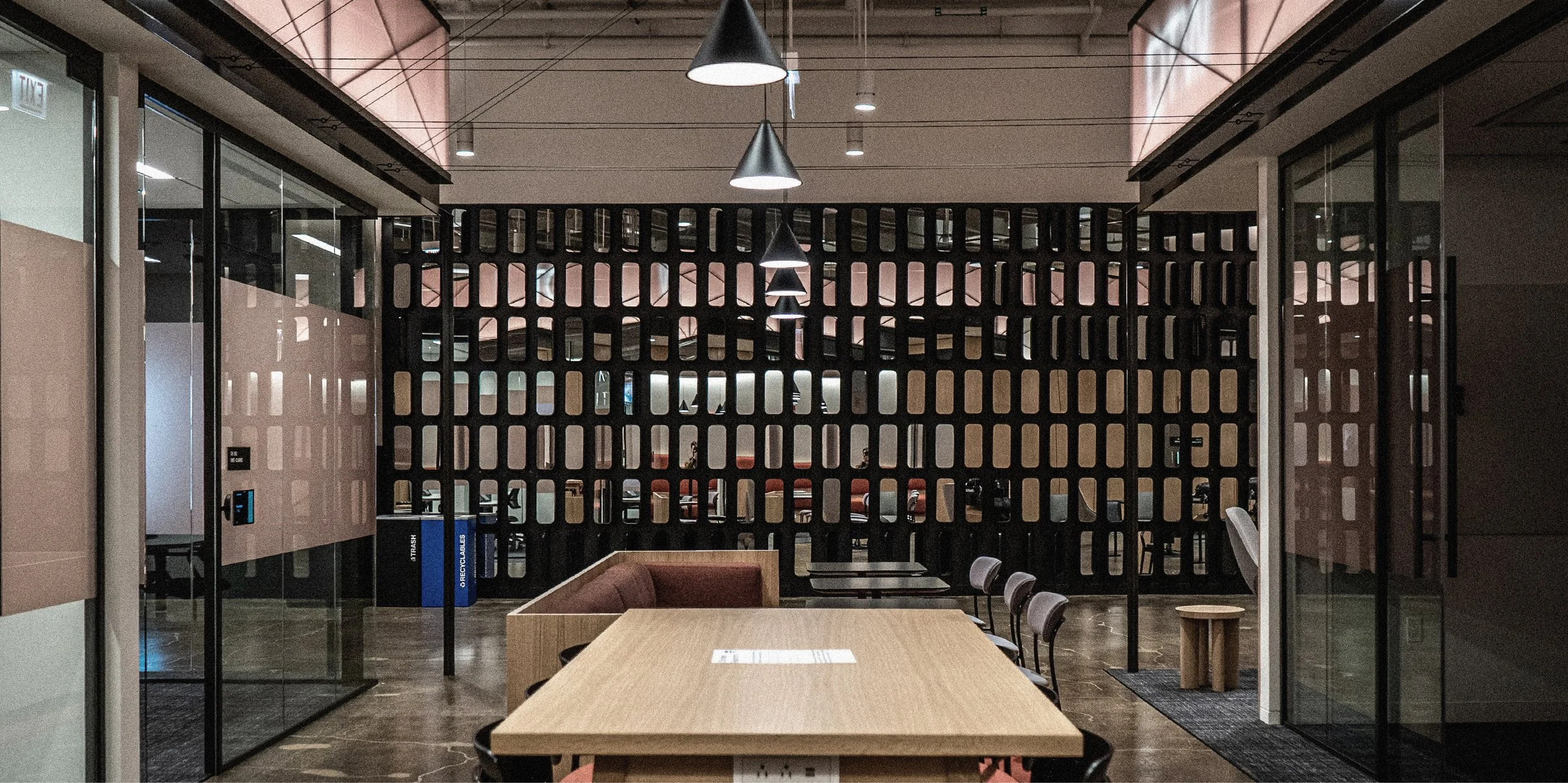 Interior of a modern office space with a wooden table, chairs, glass walls, and a decorative black lattice partition. Overhead cone-shaped pendant lights illuminate the area.