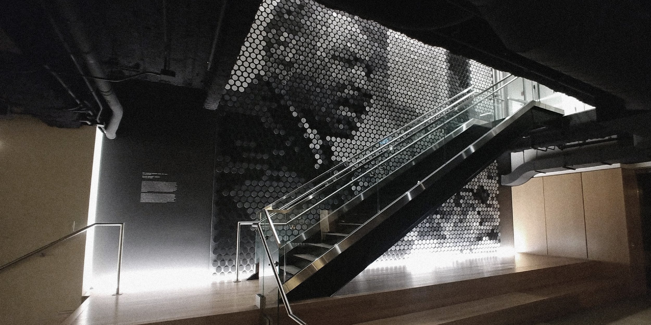 Interior of a building with a staircase, black walls, a large artistic mural made of circular dots featuring a woman's face, and subdued lighting.