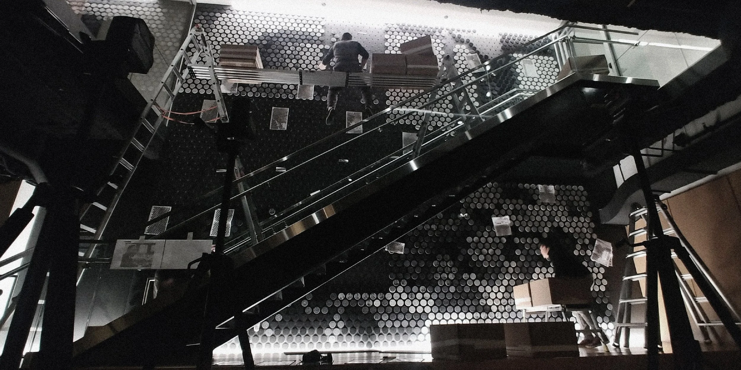 View of a modern interior space shown from below, featuring people working on a construction or setup project. There are ladders, scaffolding, and a wall with hexagonal patterns, with some furniture and equipment visible.