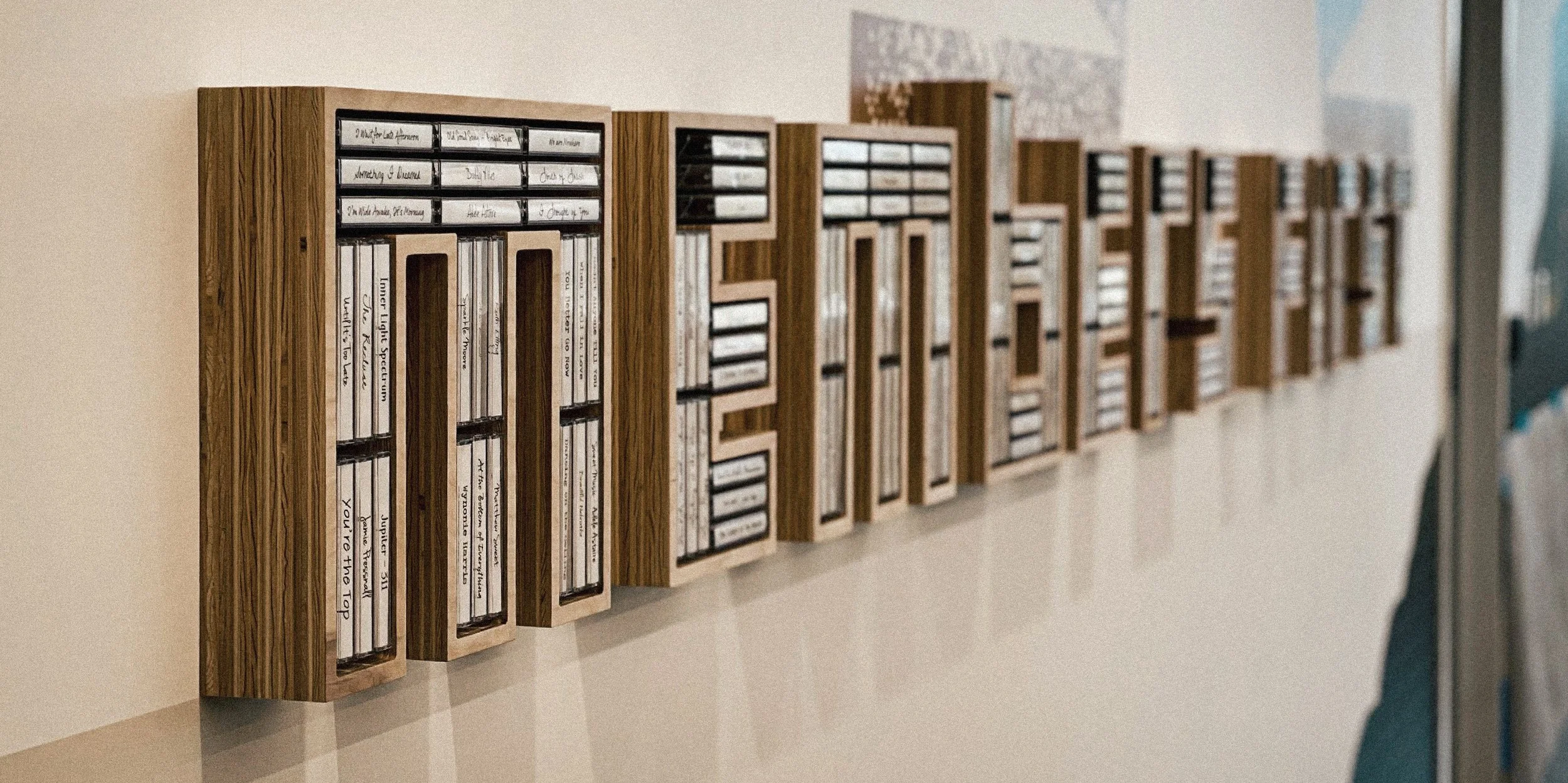 Wooden wall-mounted display cases containing cassette tapes with white labels, arranged in a row along a wall.