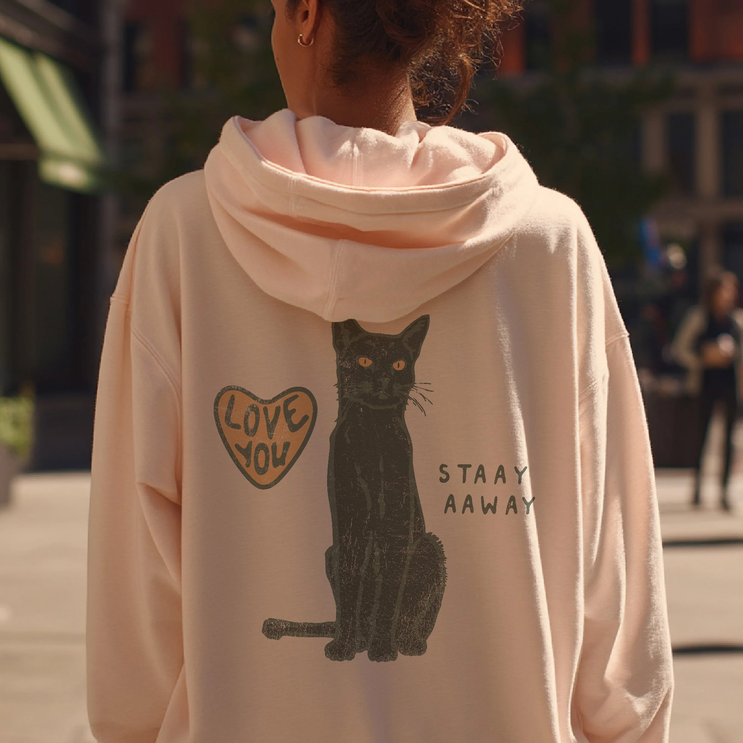 Back of a person wearing a light pink hoodie with a black cat illustration, a heart with "Love You" written inside, and the phrase "Stay Away" on the hoodie.