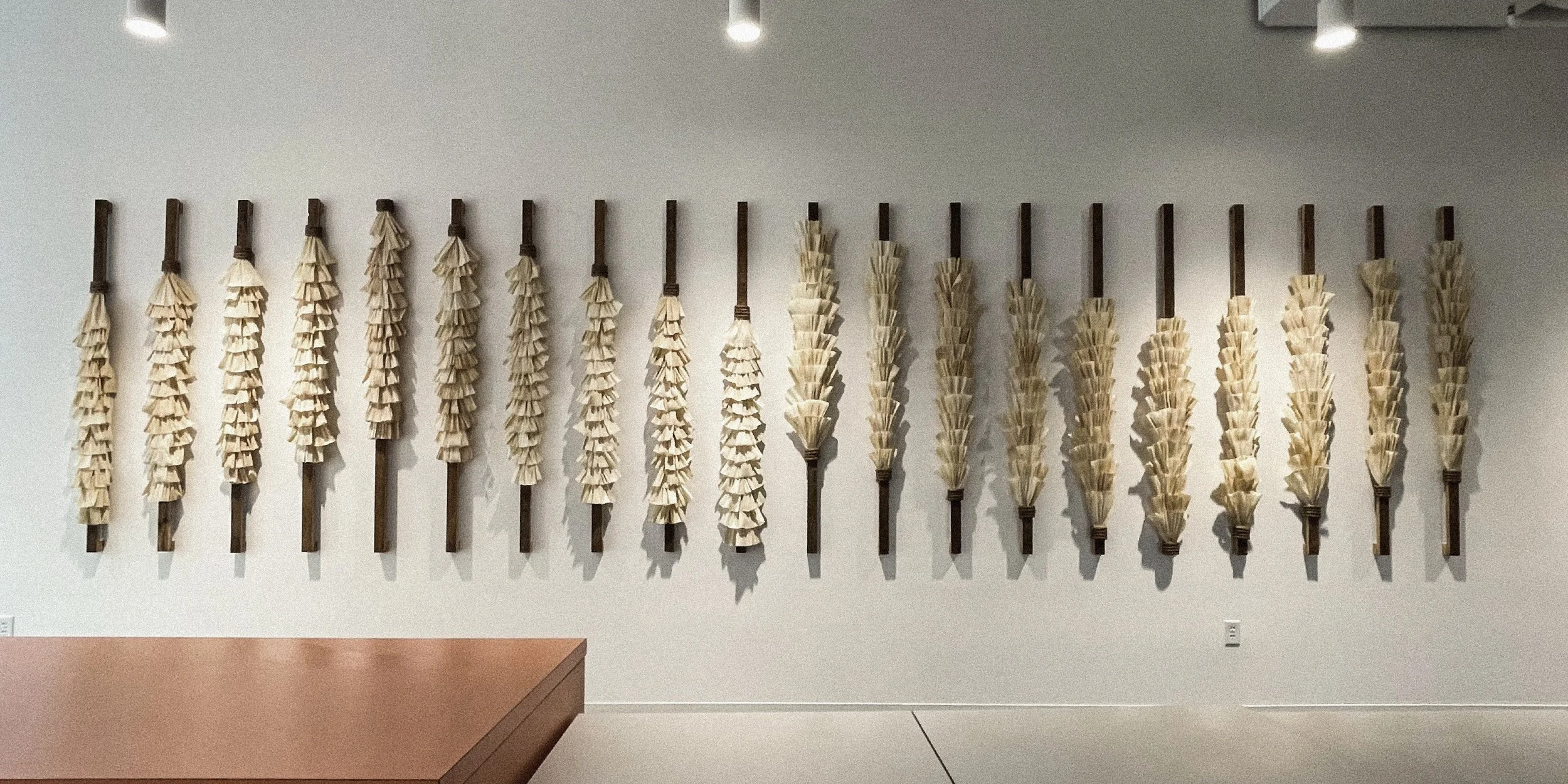 Wall display of vertical wooden sticks with layered beige paper or fabric decorations attached, arranged in a pattern.