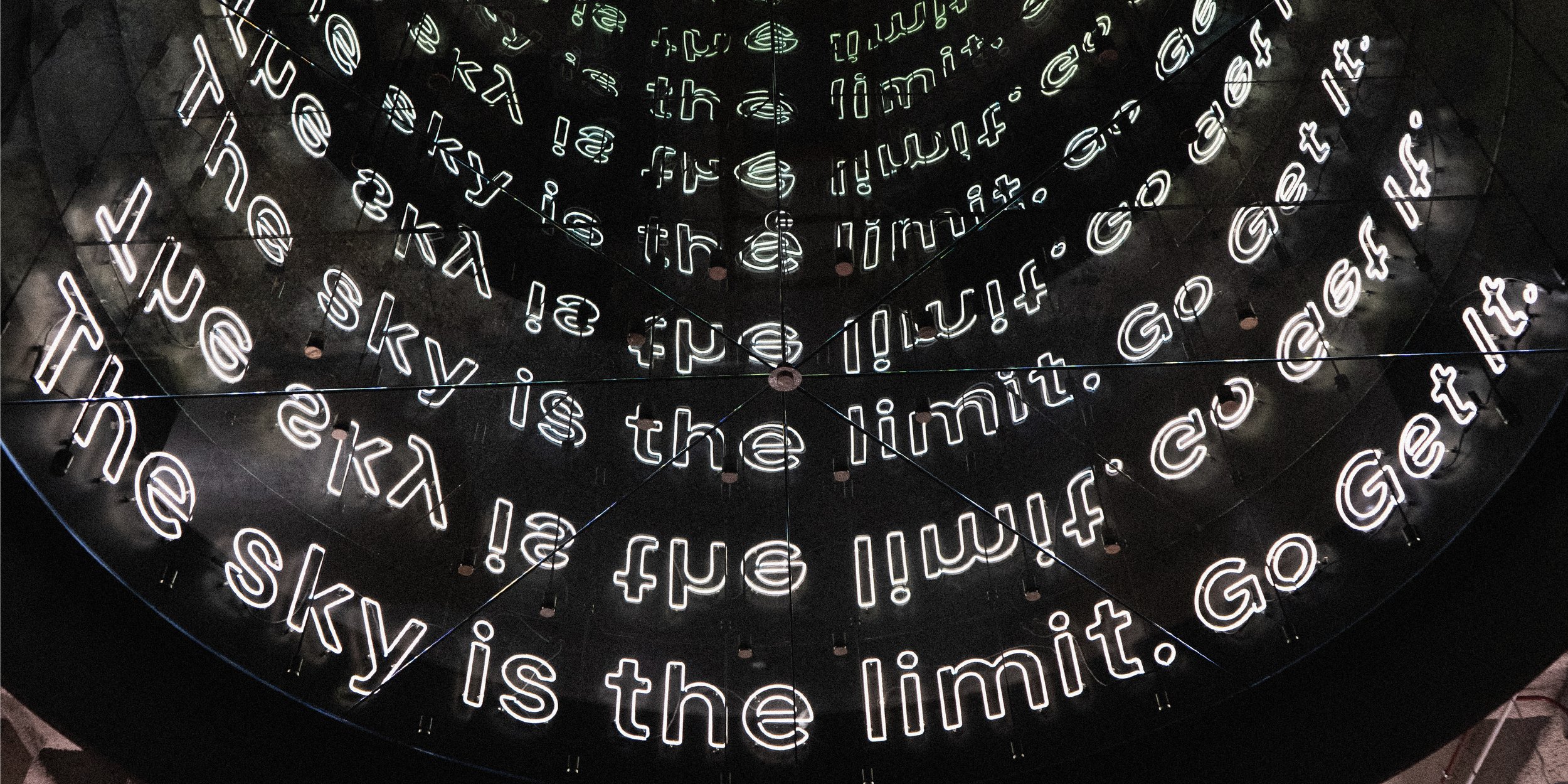 Neon sign displaying the phrase "sky is the limit!" repeated in a circular pattern with glowing white letters on a dark background.