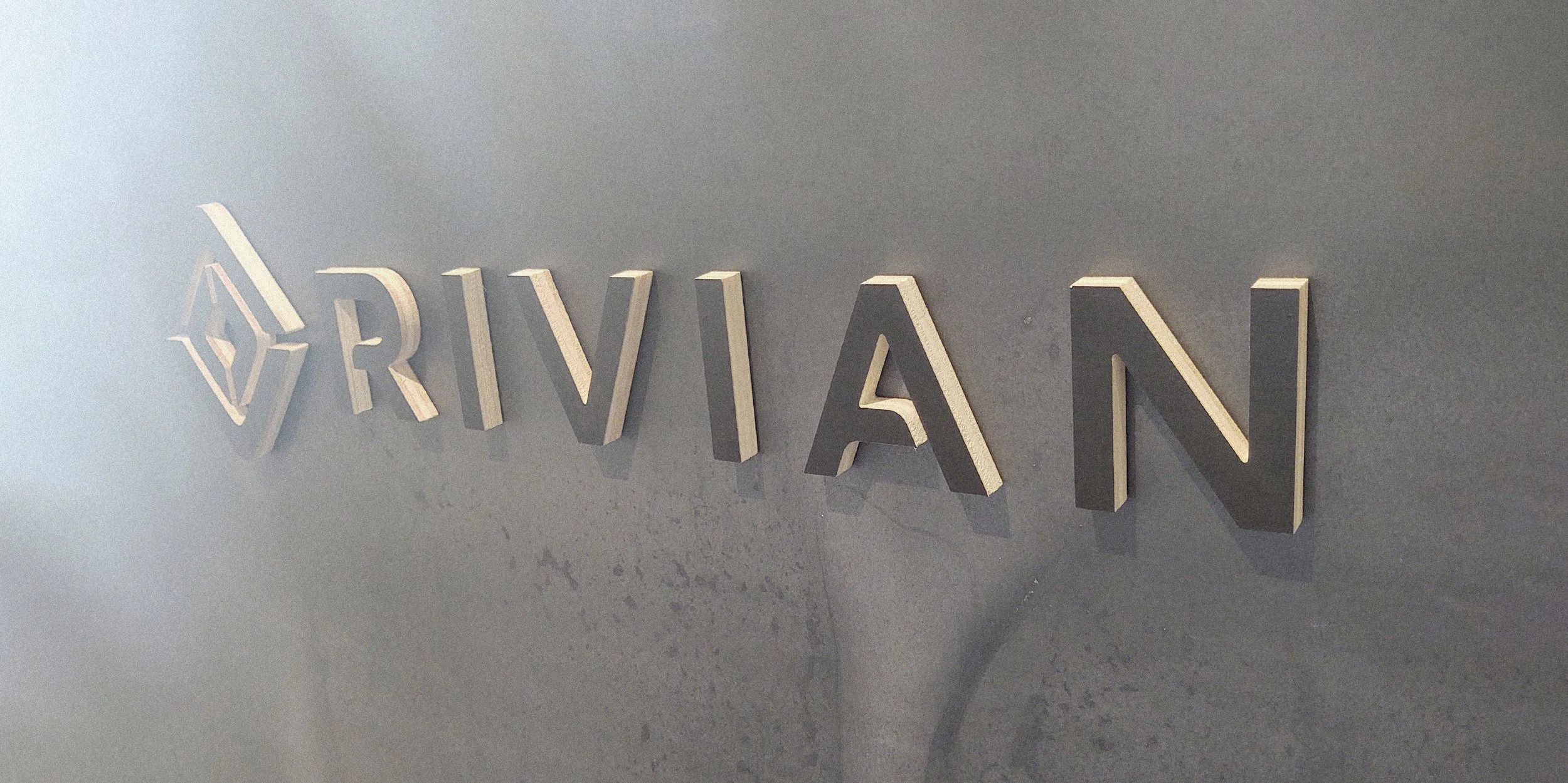 Close-up of a metallic sign that says 'RIVIAN' mounted on a grey wall.