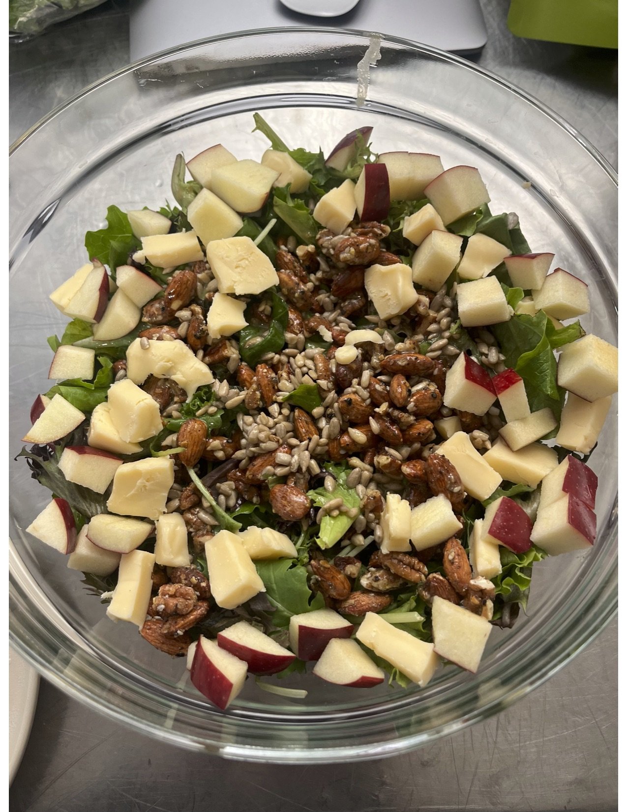 A glass bowl containing a salad with chopped red apples, mixed greens, nuts, and seeds.