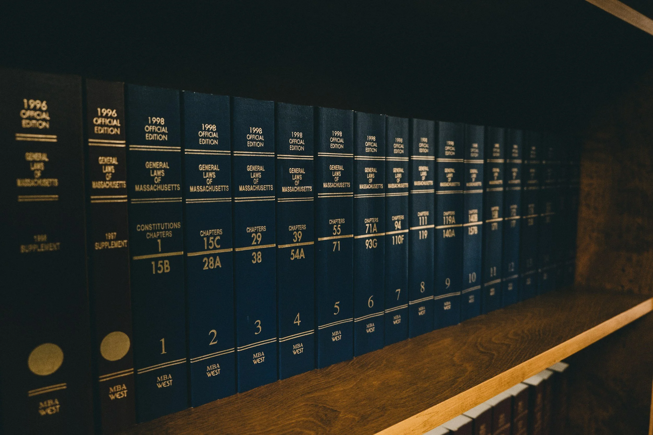 A set of blue legal books titled 'General Laws of Massachusetts' arranged on a wooden shelf, with volume numbers and chapters listed on the spines. Legal writing. 