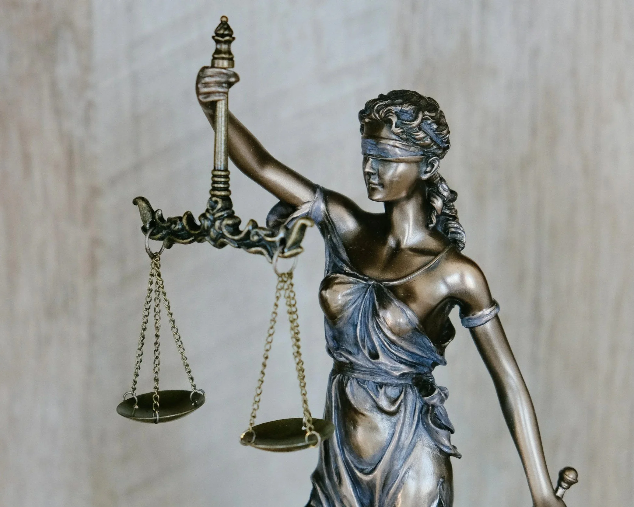 A metallic sculpture of Lady Justice, blindfolded, holding scales in her left hand and a sword in her right hand, standing against a neutral background. Showcasing neutrality. 
