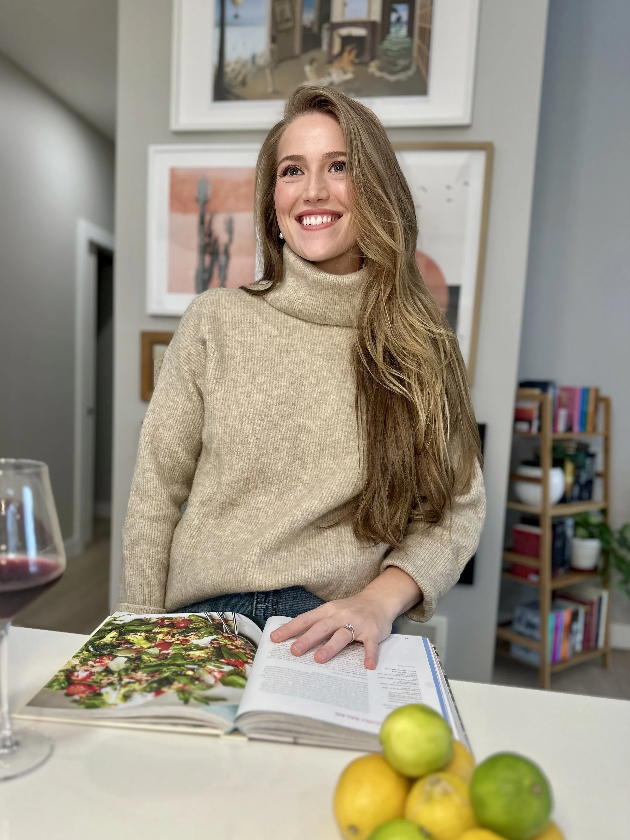 A young woman with long blonde hair smiling in a beige turtleneck sweater in a home setting, with open cookbook, lemons, and a glass of red wine on the table.