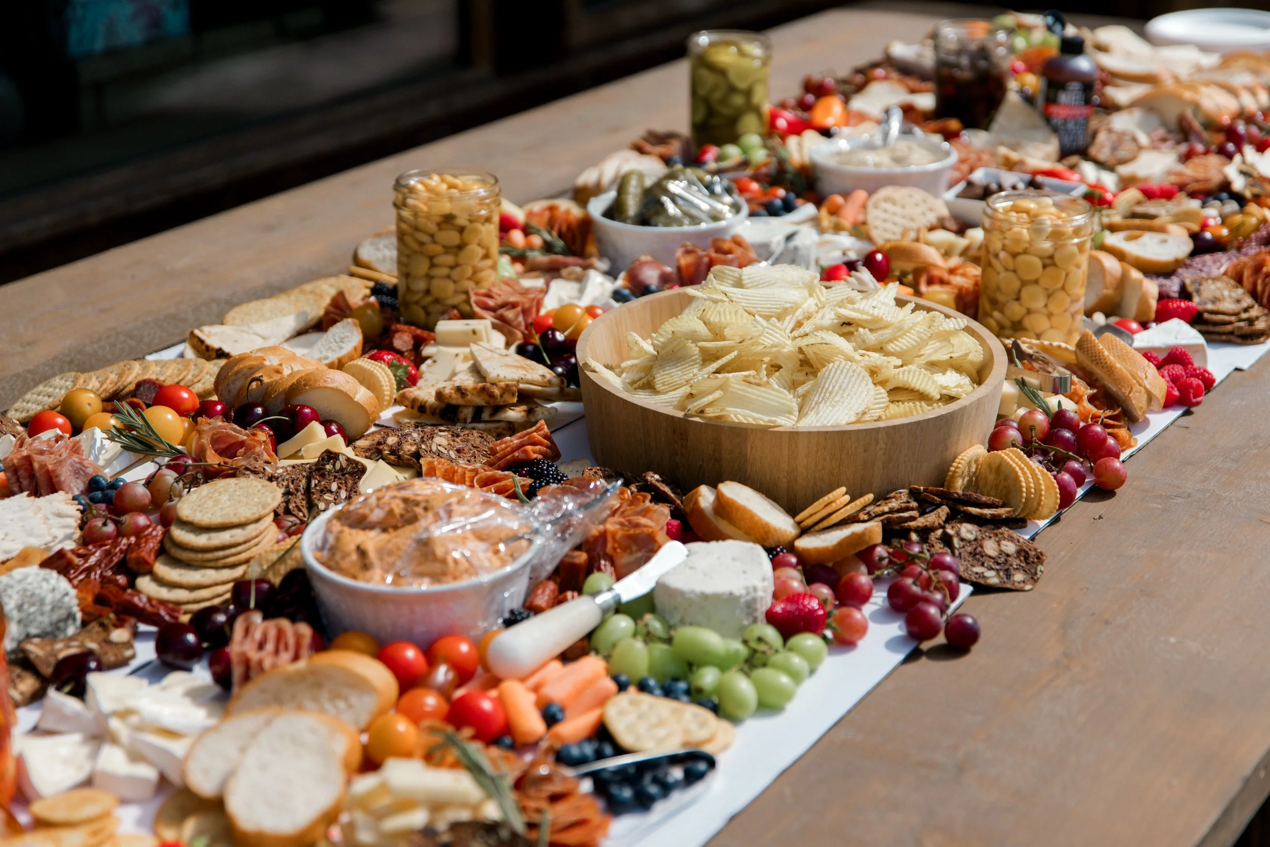 A long table filled with an assortment of cheeses, crackers, bread, grapes, cherry tomatoes, olives, and other finger foods, likely prepared for a large gathering or party.