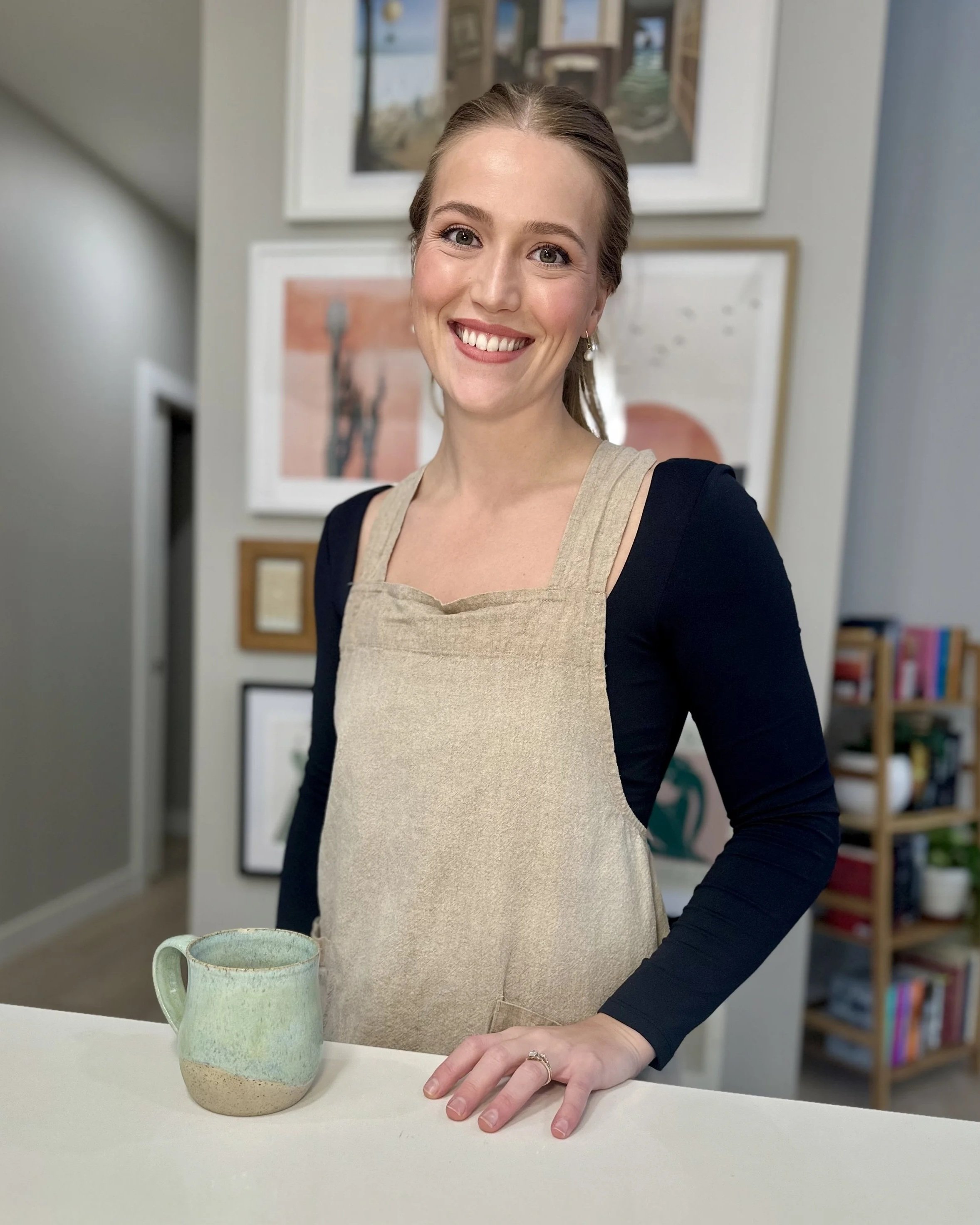 A woman with light brown hair smiling, wearing a beige apron over a navy blue long sleeve shirt, standing behind a white counter with a green ceramic mug, in a room with framed pictures on the wall and a bookshelf in the background.