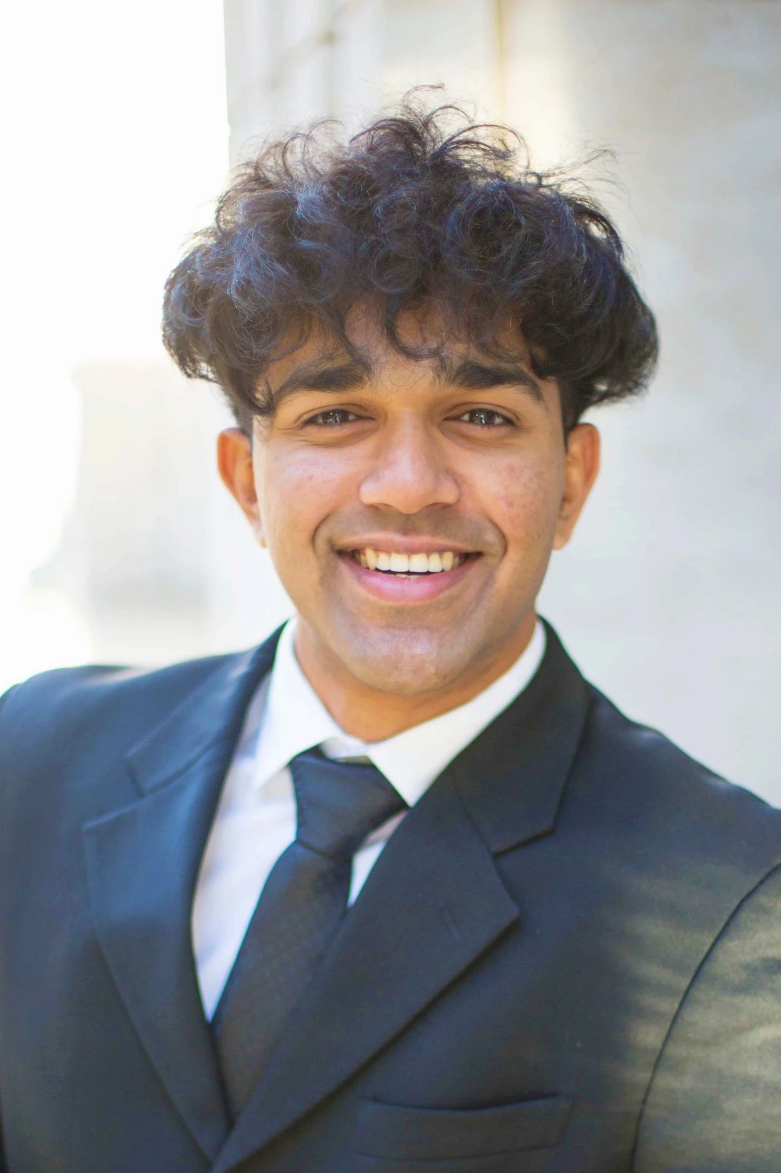 A young man with curly dark hair wearing a black suit, white shirt, and black tie, smiling outdoors. Sabin Alam- the founding principal