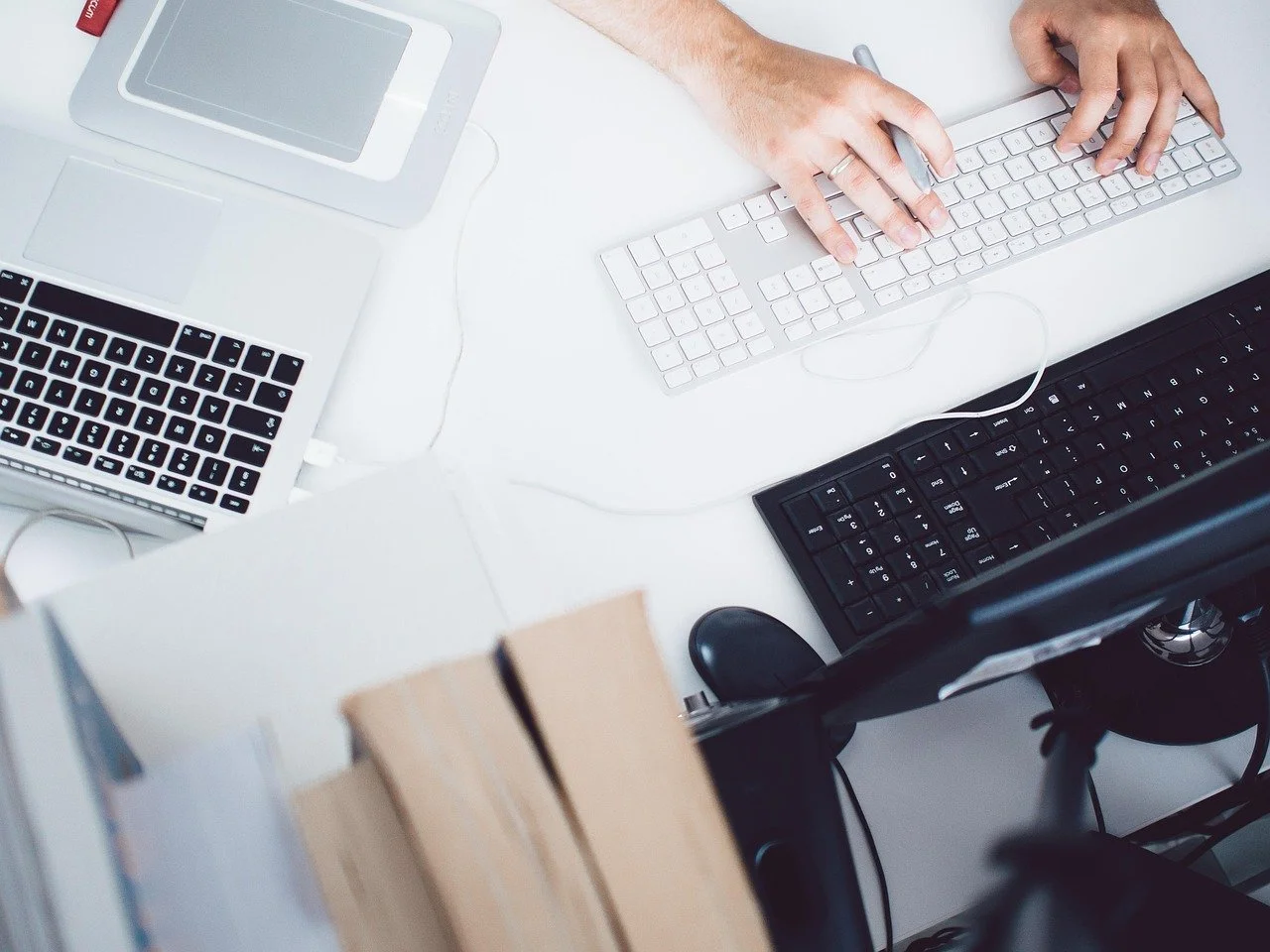 Top-down view of a white desk with a person's hands using a wireless keyboard and mouse, a laptop, a phone, and other office supplies.