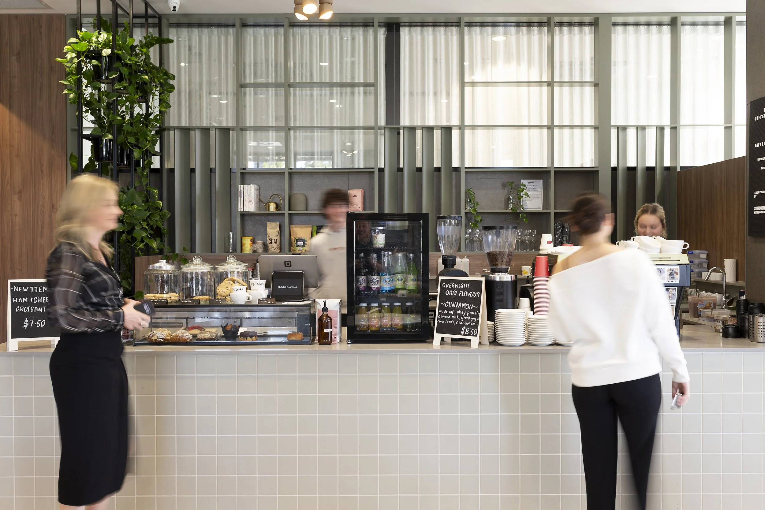 Interior of a modern coffee shop with a counter, menu signs, two female customers, and staff working behind the counter.