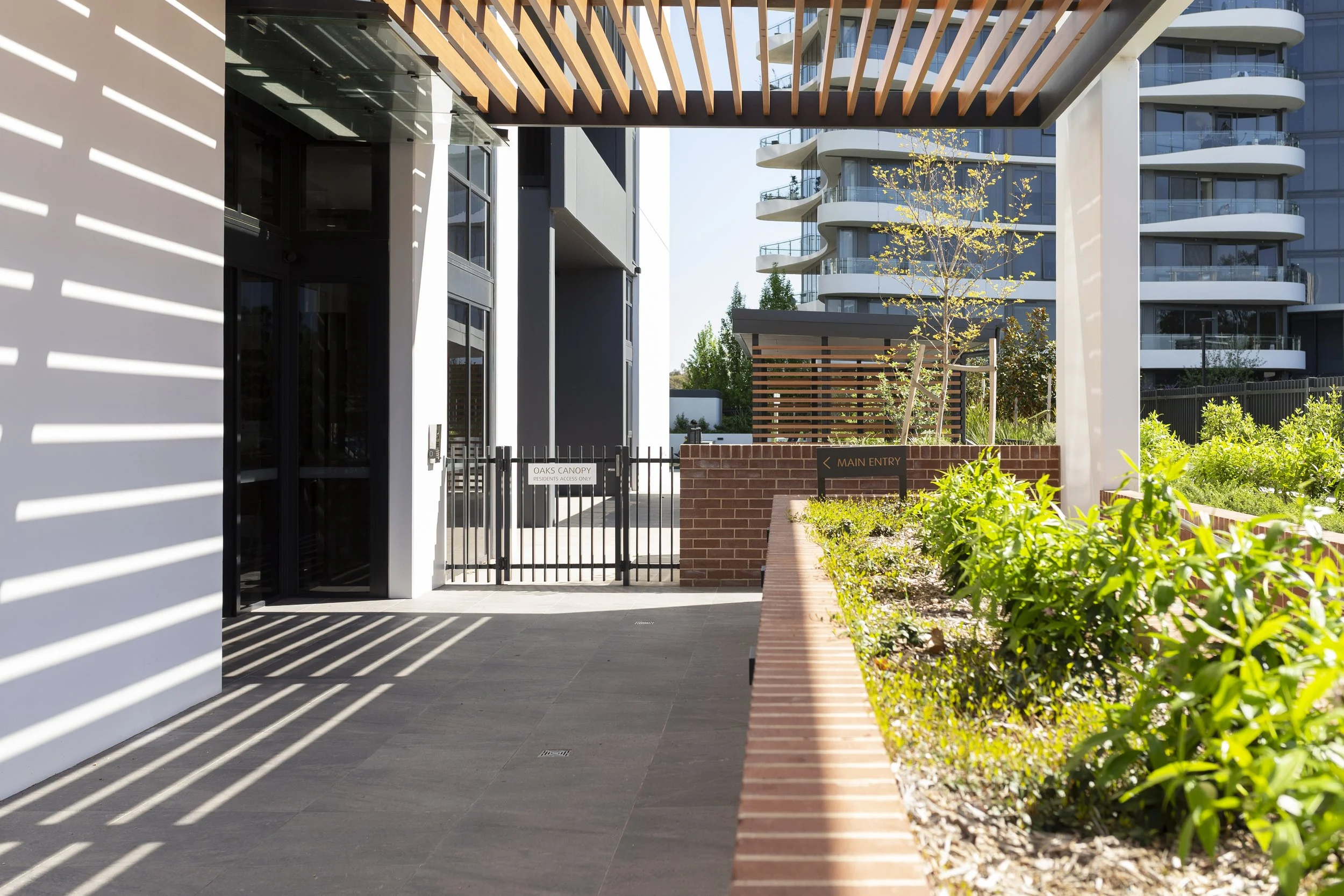 Entrance area of a modern apartment complex with a gated entry and landscaped garden, featuring a sign that says 'Main Entry' and another sign that reads 'Oaks Canopy'.