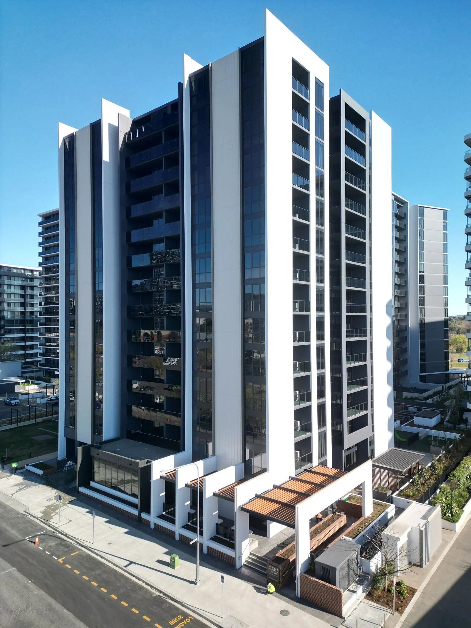 Modern multi-story residential building with glass balconies and white exterior, located on a city street with clear blue sky in background.