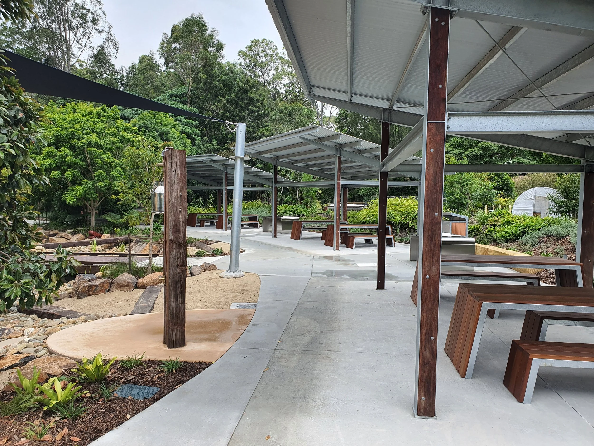 Outdoor seating area with shaded structures, wooden benches, and surrounding greenery.