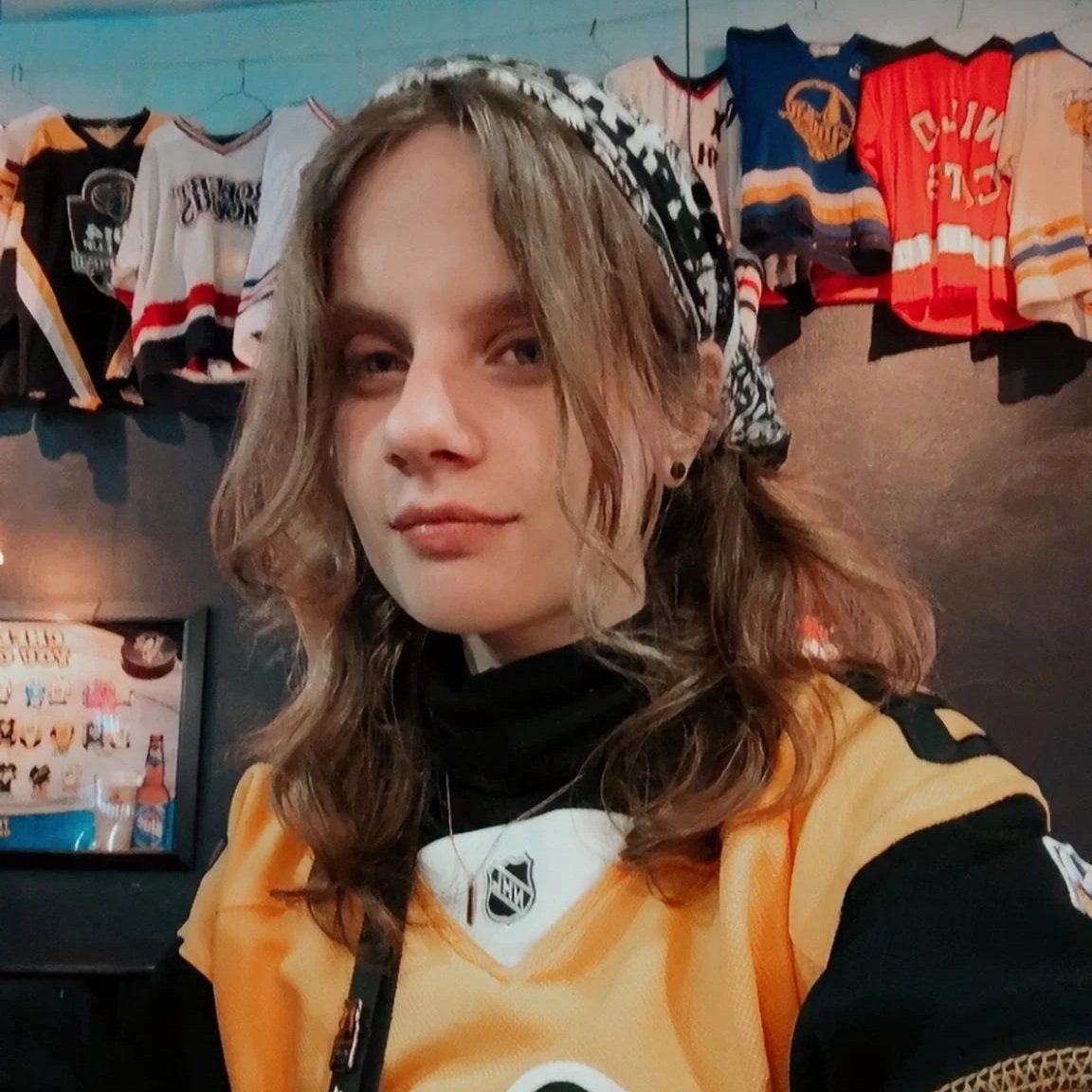 A woman with wavy brown hair and earrings taking a selfie inside a hockey fan shop, wearing a Nashville Predators jersey with NHL logo, with hockey jerseys hanging on the wall behind her.