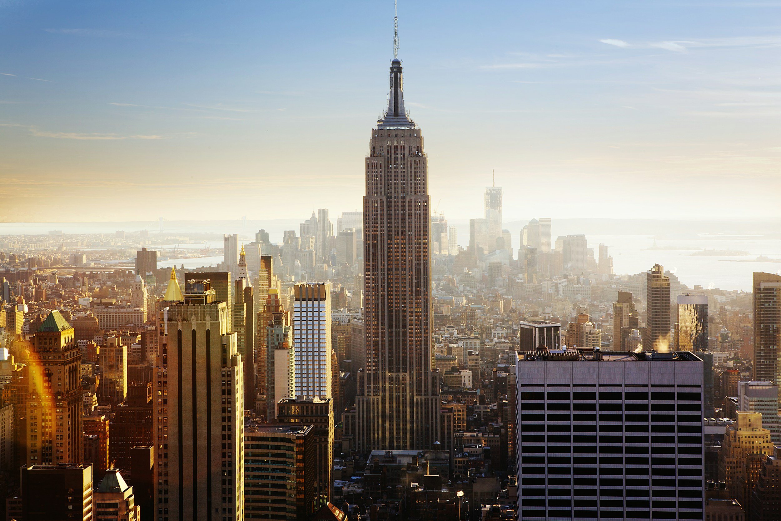 A city skyline featuring the Empire State Building, with other skyscrapers and buildings, under a partly cloudy sky during sunset.