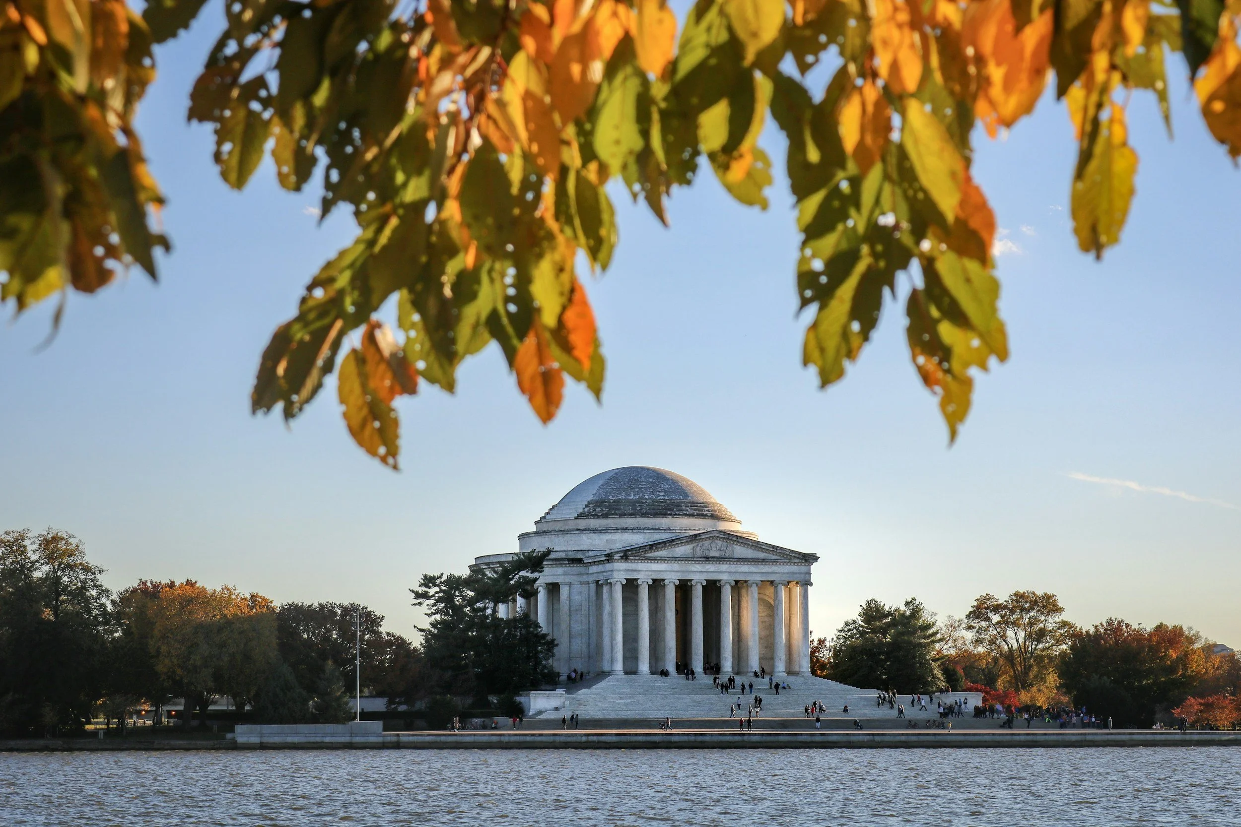 The Thomas Jefferson Memorial in Washington, D.C., seen from across the water with autumn leaves in the foreground.