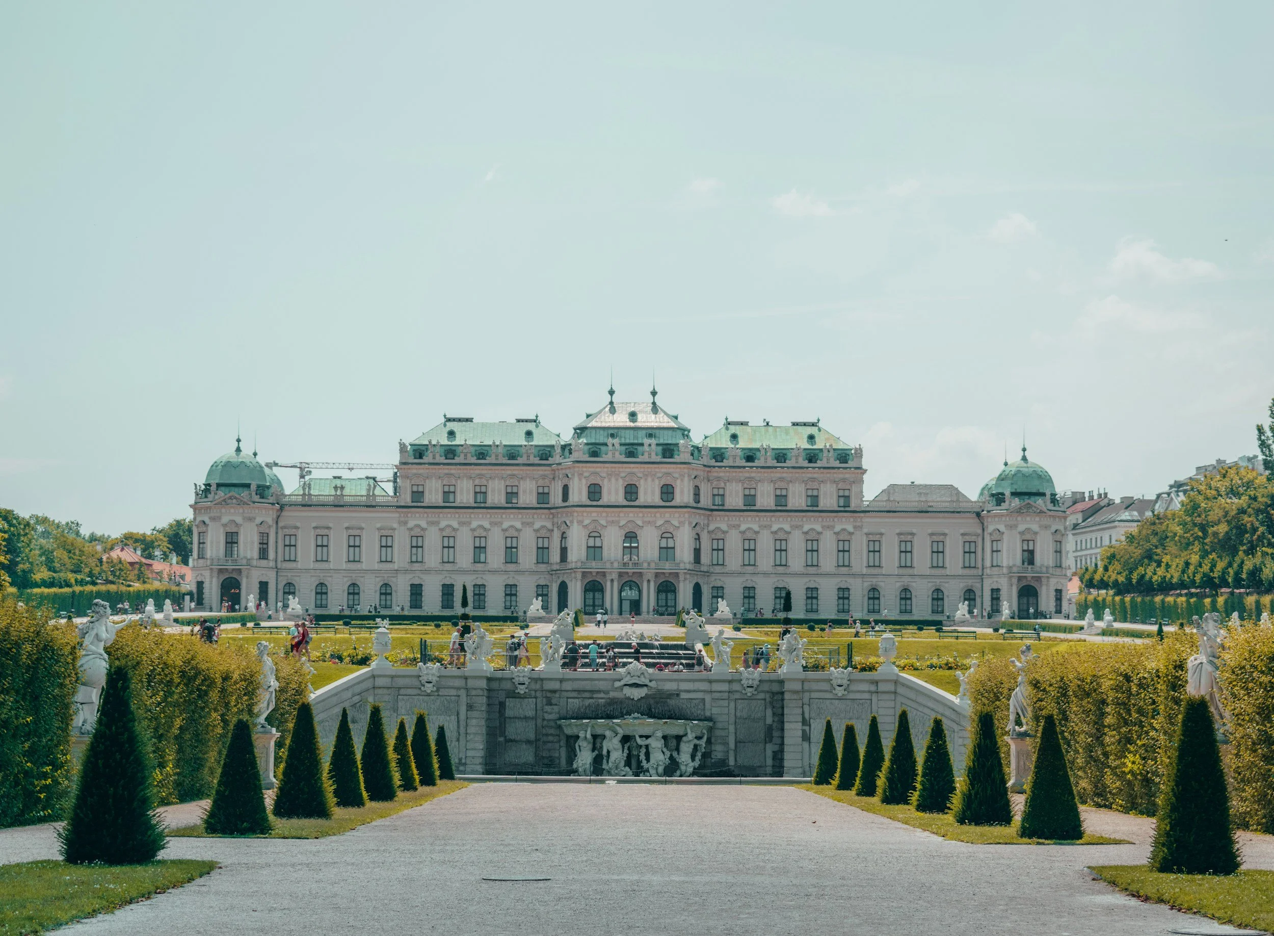 A grand, historic palace with ornate architecture, surrounded by a manicured garden and statues, under a clear sky.