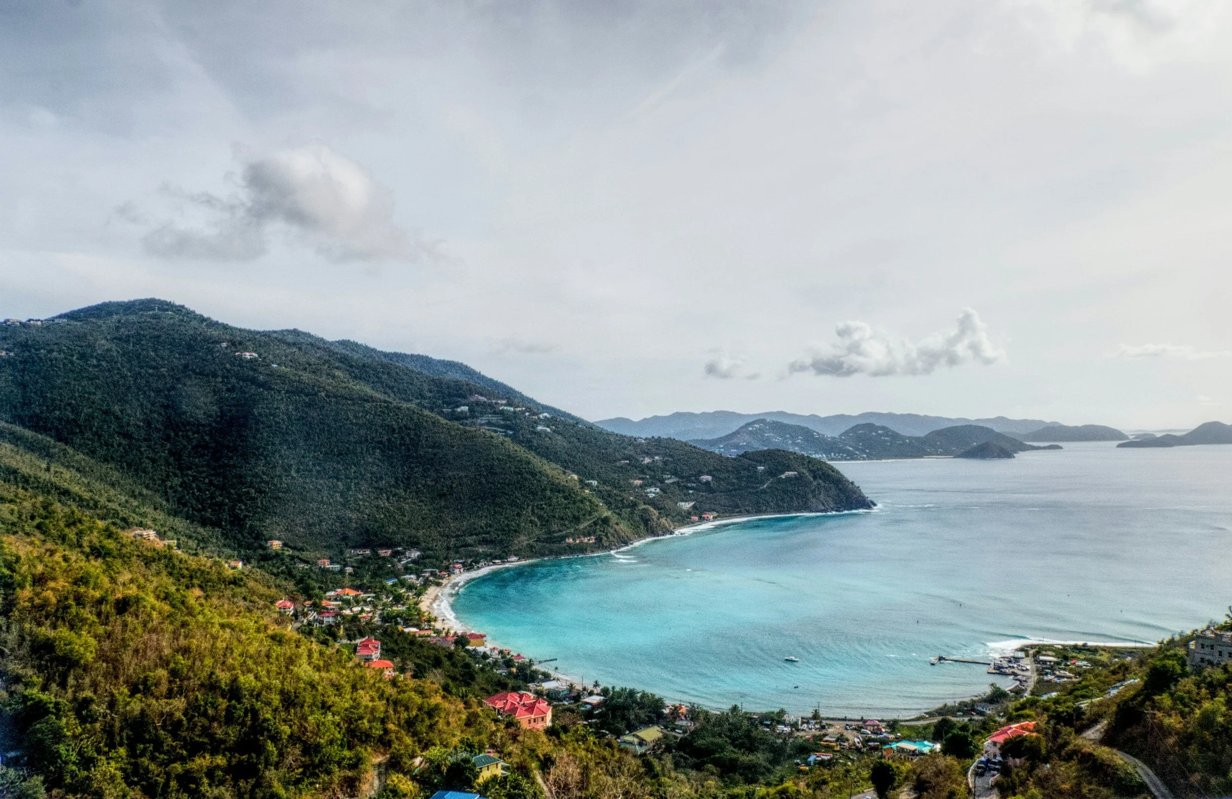 Scenic view of a coastal bay with a crescent-shaped beach, surrounded by green hills and a small town, under a partly cloudy sky.