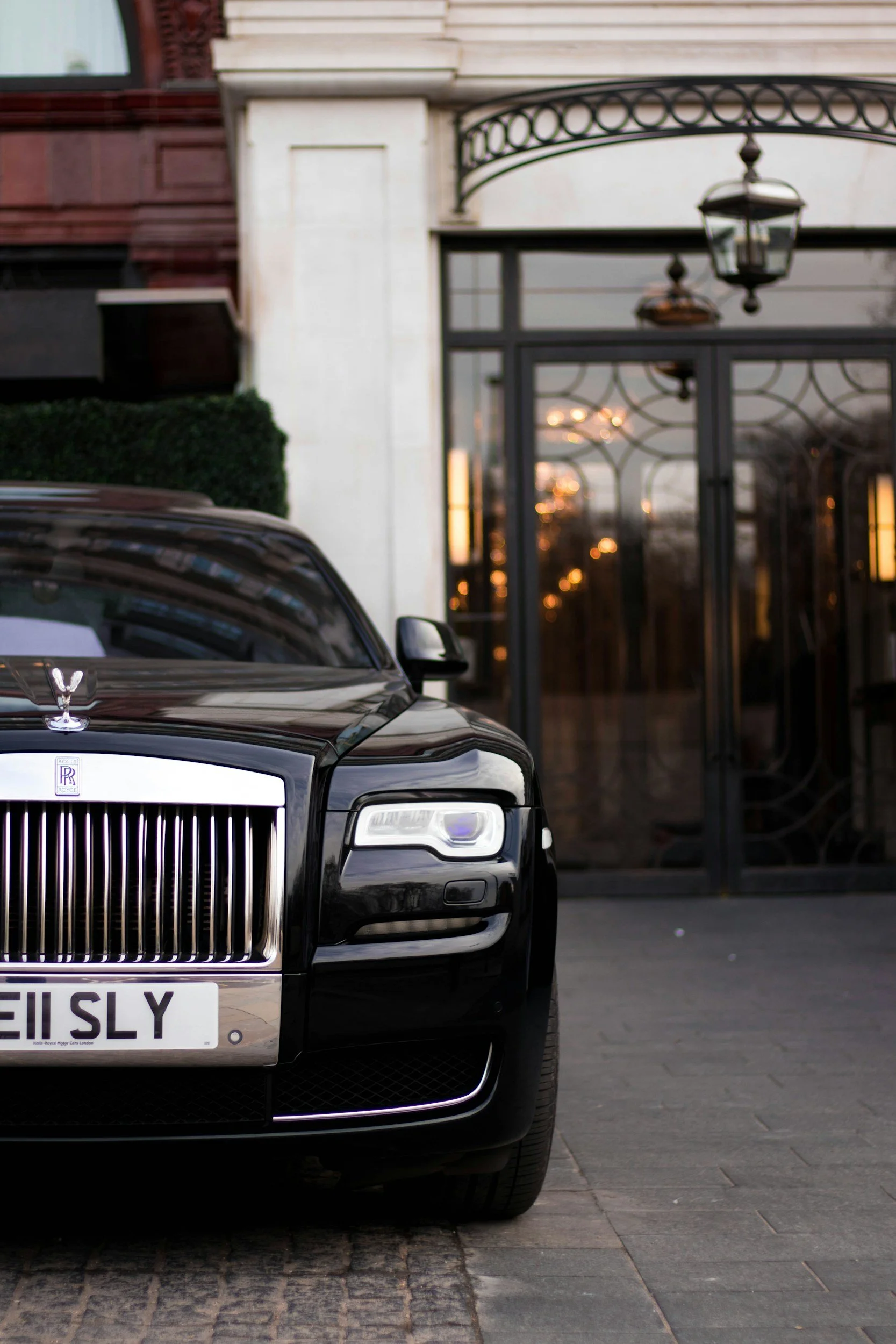 Black Rolls-Royce car parked outside an elegant building with glass doors and lanterns, during sunset.