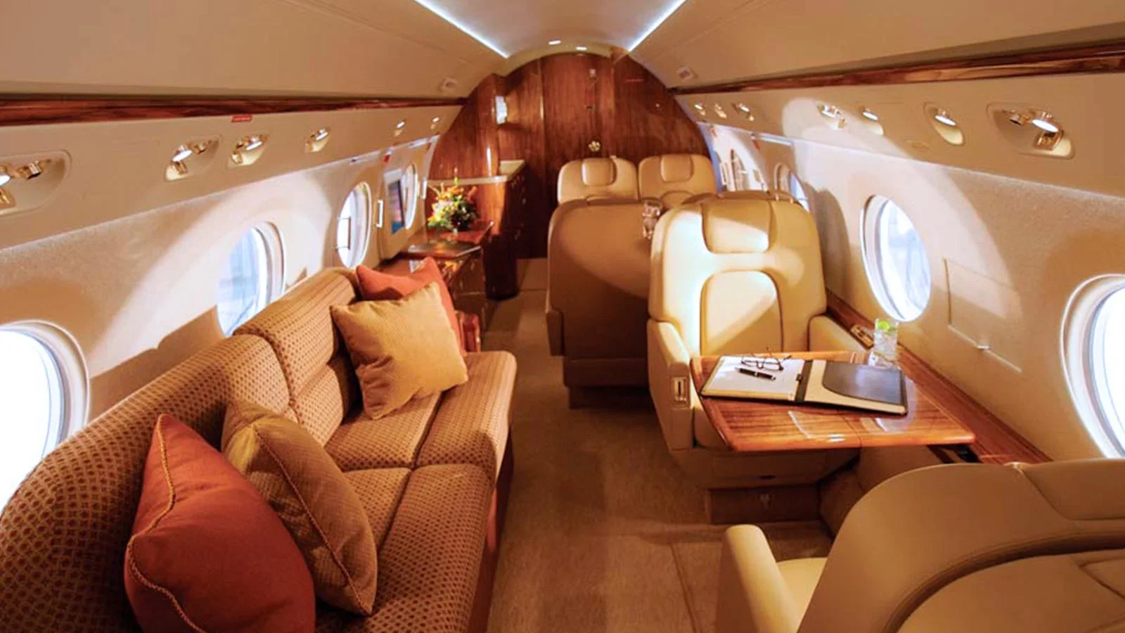 Interior of a private jet with beige leather seats, a sofa with cushions, and wooden accents. There are windows along the sides and a table with a folder, pen, and water bottle on it.