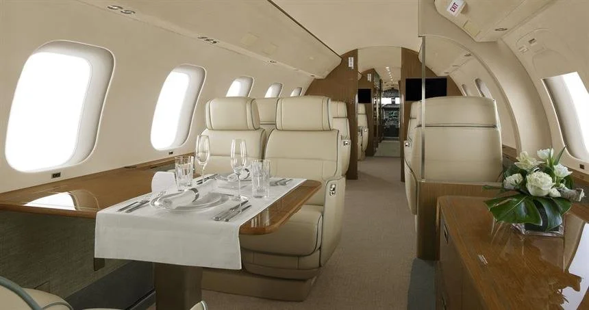 Inside of a private jet with a dining table set with wine glasses, plates, and utensils, surrounded by beige leather seats, with windows along the cabin walls.