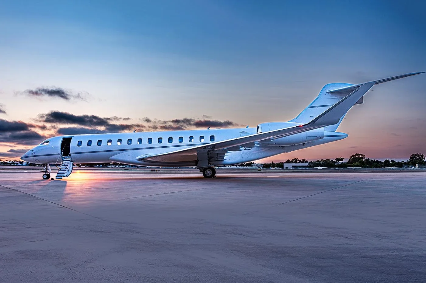 A sleek, white private jet parked on an airport tarmac at sunset, with stairs leading into the aircraft and low clouds in the sky.