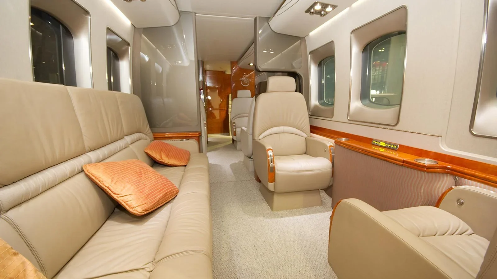 The interior of a private jet with beige leather chairs and couches, orange pillows, and wooden accents along the walls.