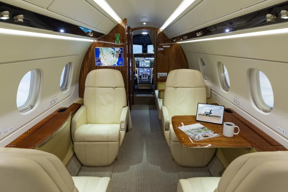 Inside the cabin of a private jet with beige leather seats, wooden accents, windows, and a small table with a tablet, coffee mug, and magazines.