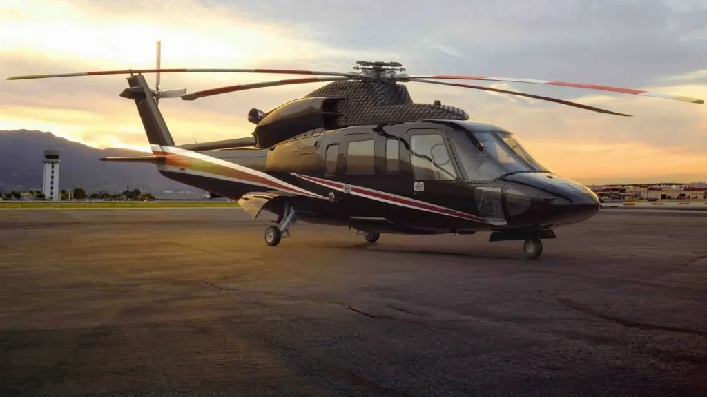 A black and red helicopter parked on an airport tarmac at sunset, with mountains and a control tower in the background.