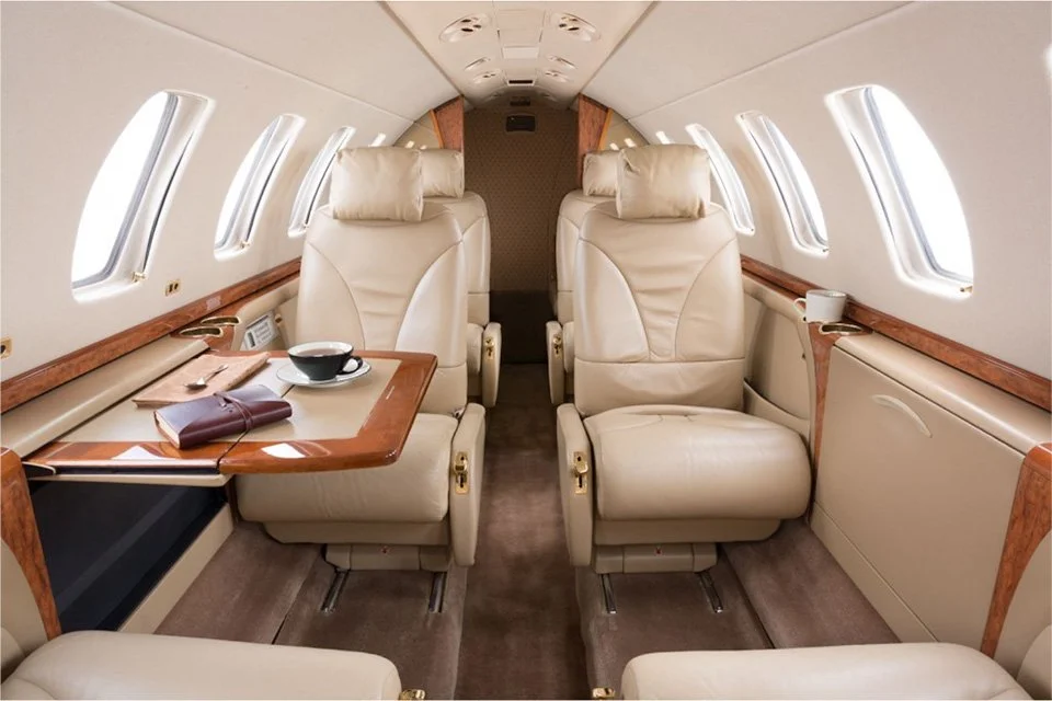 Interior of a private jet with leather seats, small windows, wooden accents, and a small table with a cup and book.