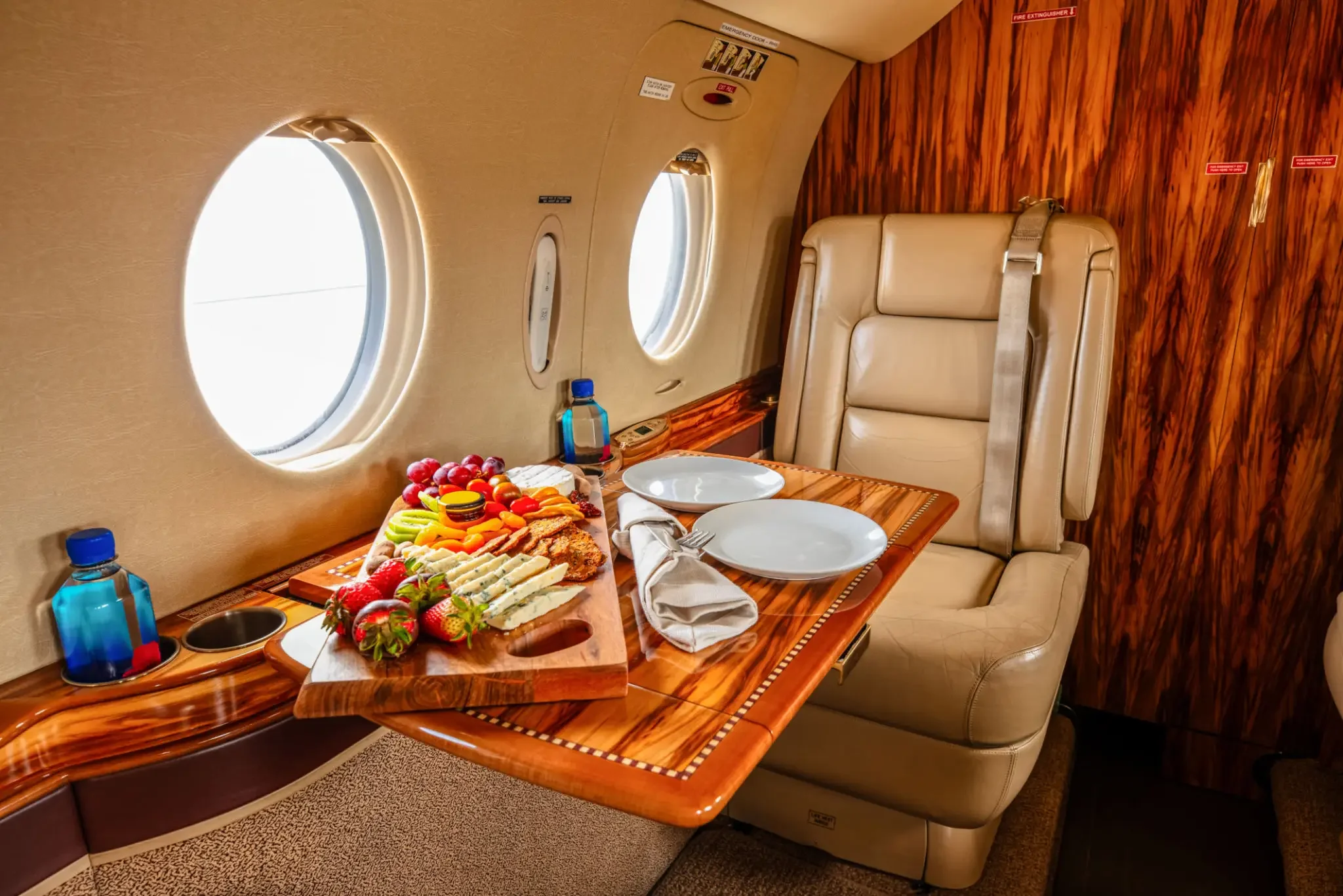 A luxurious airplane interior with a wooden table set with plates, napkins, and cutlery, and a tray of assorted fruits, cheeses, and crackers. The seating is cream-colored leather, with a large window showing the sky outside.