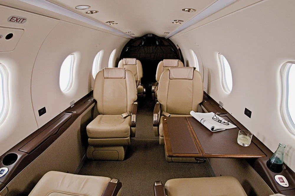 Interior of a private jet with beige leather seats, a wooden table with glasses and papers, and windows along the side.