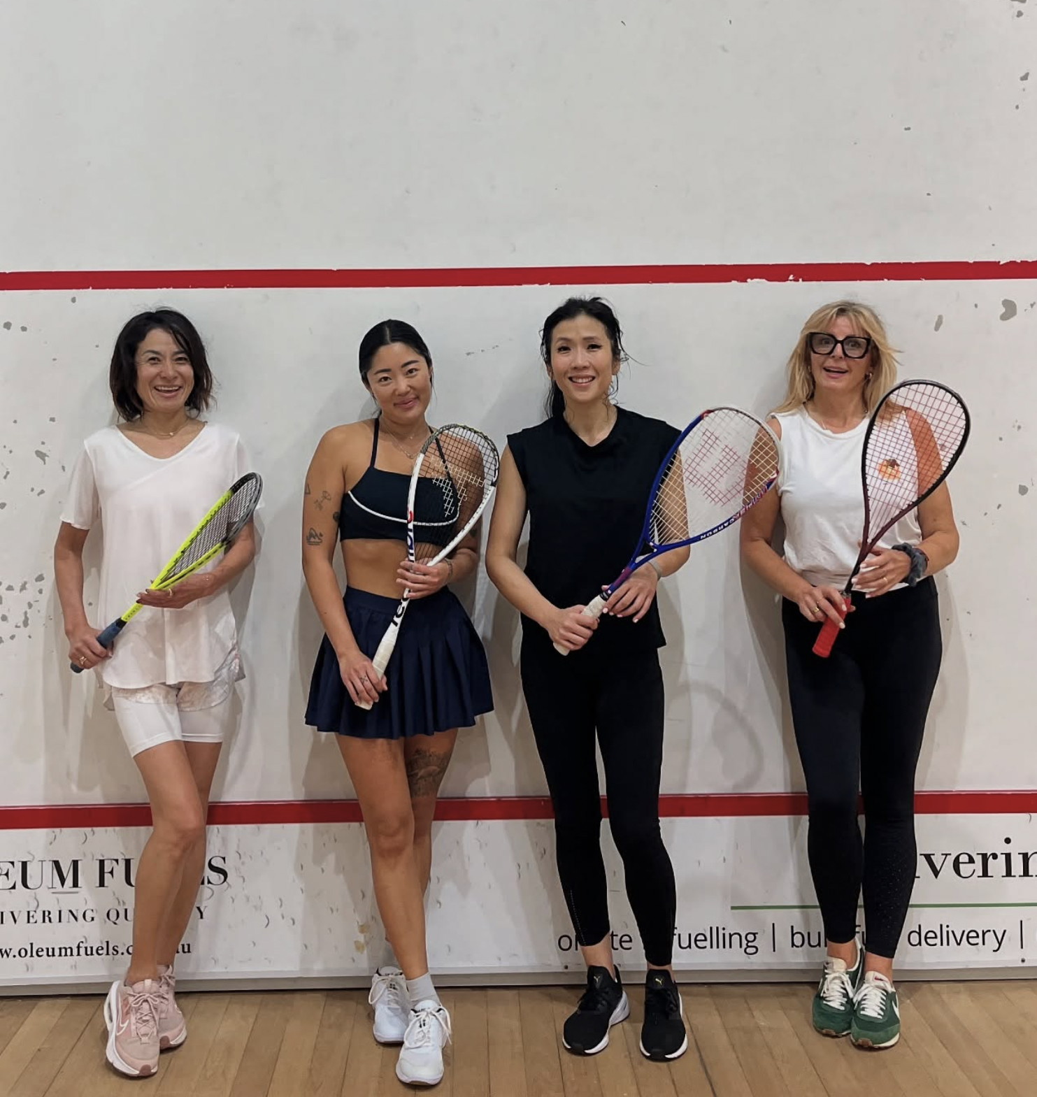 Four women standing in a squash court holding squash rackets, dressed in athletic or casual sportswear, smiling at the camera.