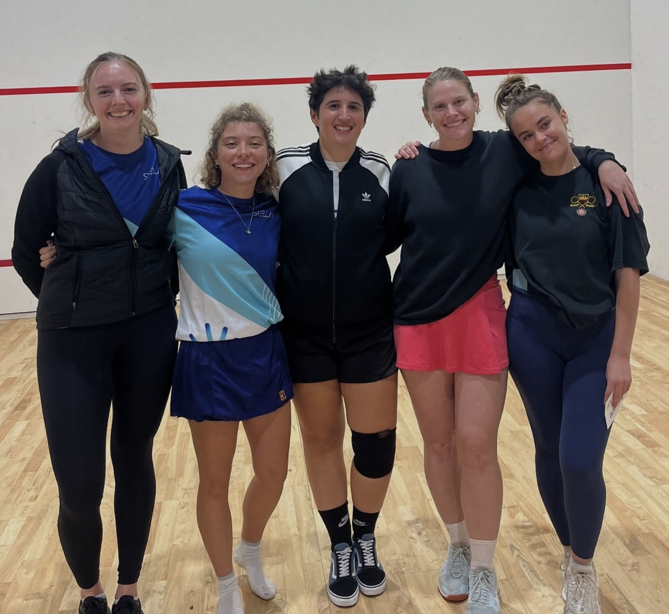 Five young women standing together on a wooden floor in front of a white wall with red lines, smiling and wearing sportswear.