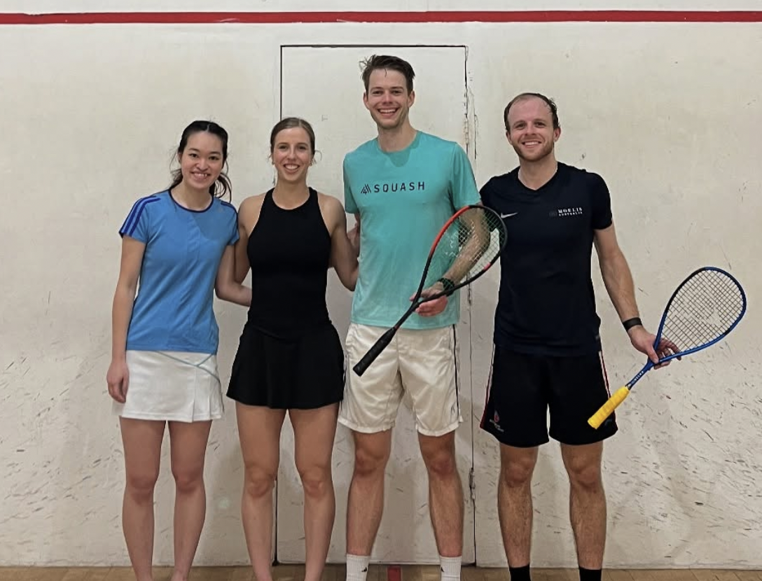 Four people standing together in front of a wall with a small door or hatch, holding squash rackets, smiling after a game.
