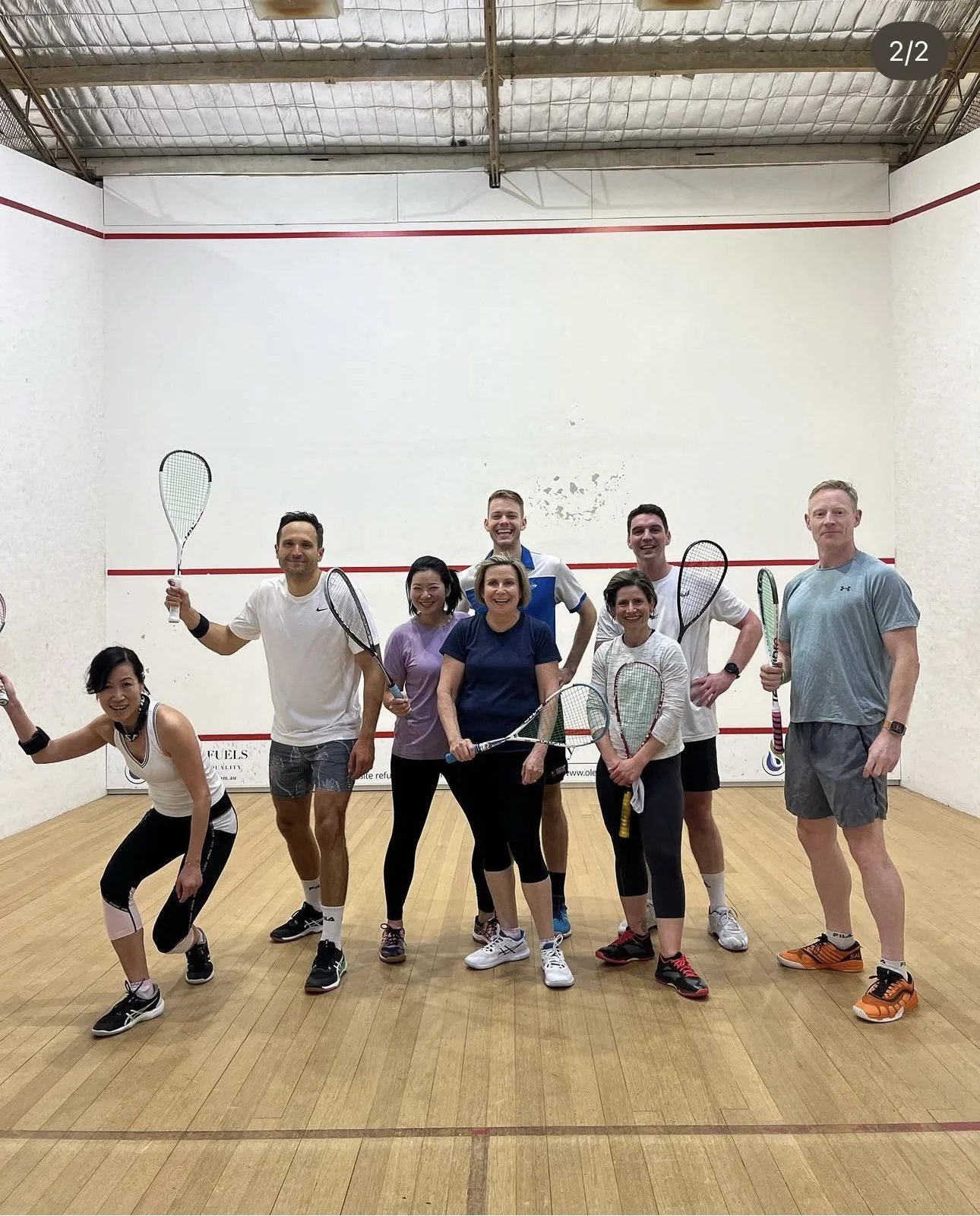 A group of nine people, 5 women and 4 men, standing with tennis rackets inside a squash court, posing for a group photo. They are smiling and dressed in sportswear.