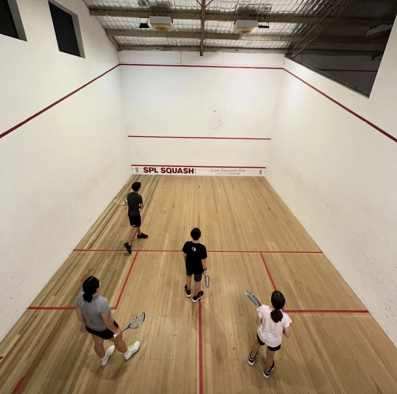 Four young people are playing squash in an indoor court with white walls and wooden flooring. They are holding rackets, and one is on the court while others wait or prepare to play.