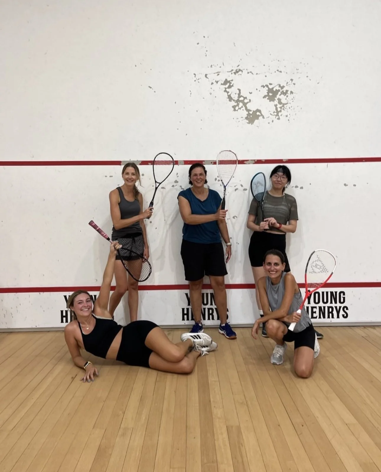Five women posing with tennis rackets on an indoor squash court. Two are standing, three are kneeling or lying on the floor. The court has a white wall with a red boundary line and the words "YOUNG HENRYS" printed on it.