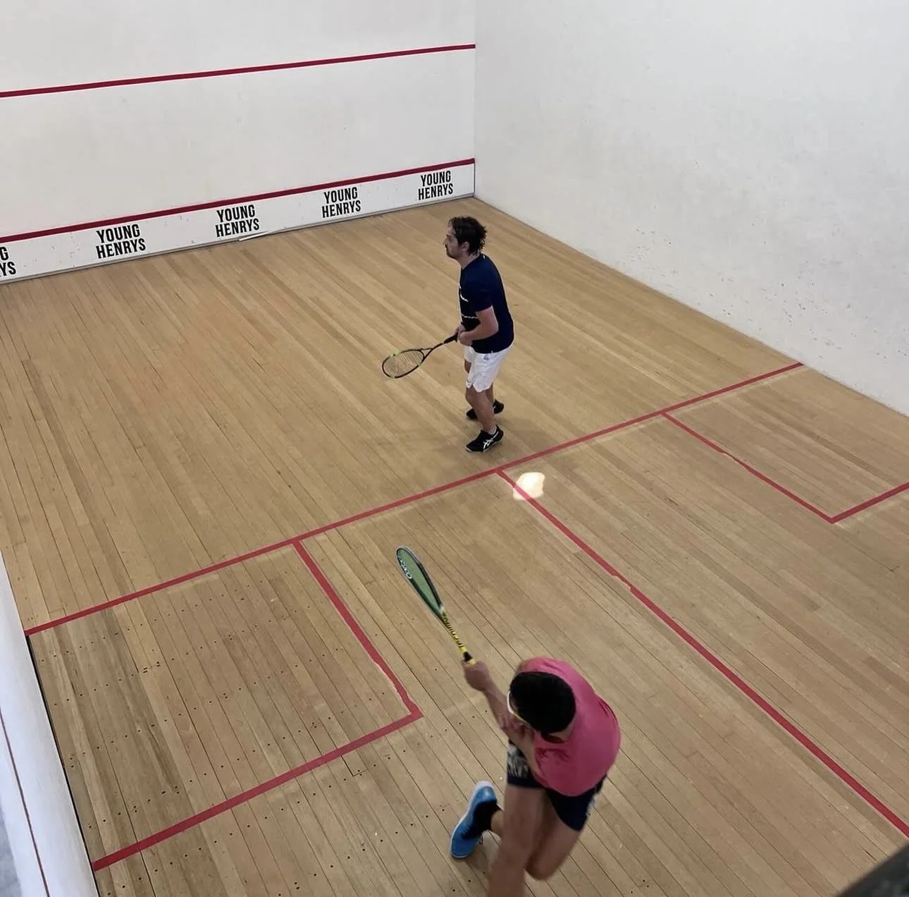 Two men playing squash on a court with wooden flooring and red boundary lines.