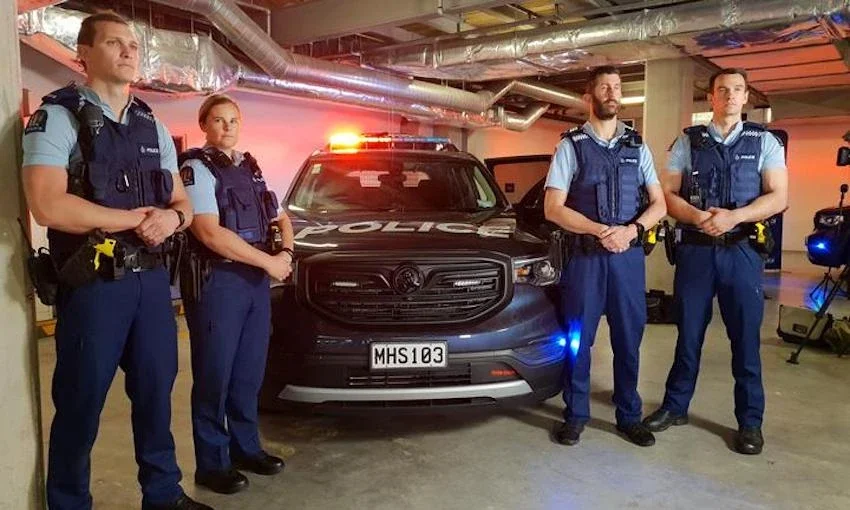 Four police officers in uniform standing in front of a police vehicle with flashing lights in an underground parking garage.