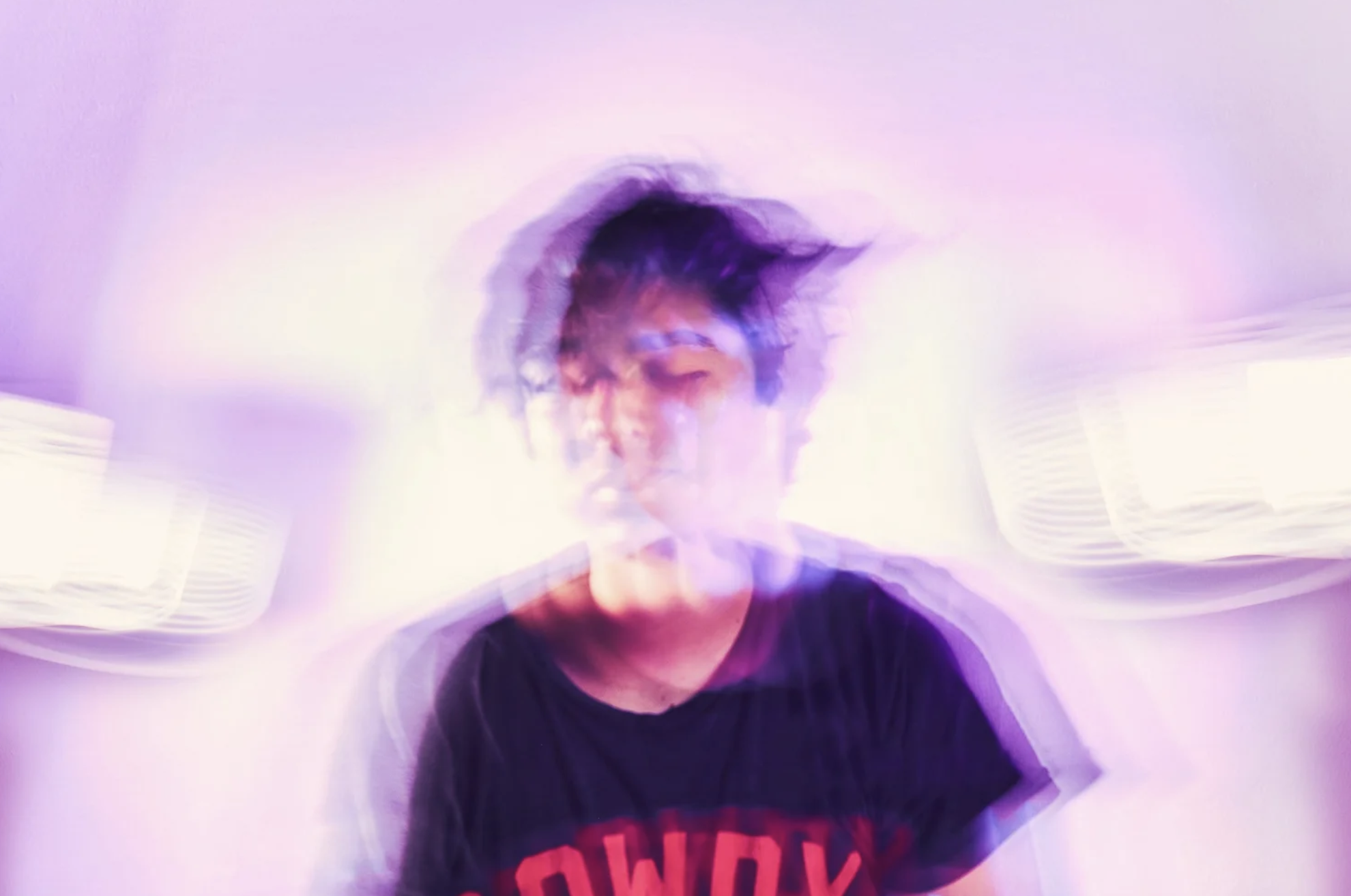 A person with dark hair and a black shirt standing against a light purple background, with a motion blur effect creating multiple overlapping images.