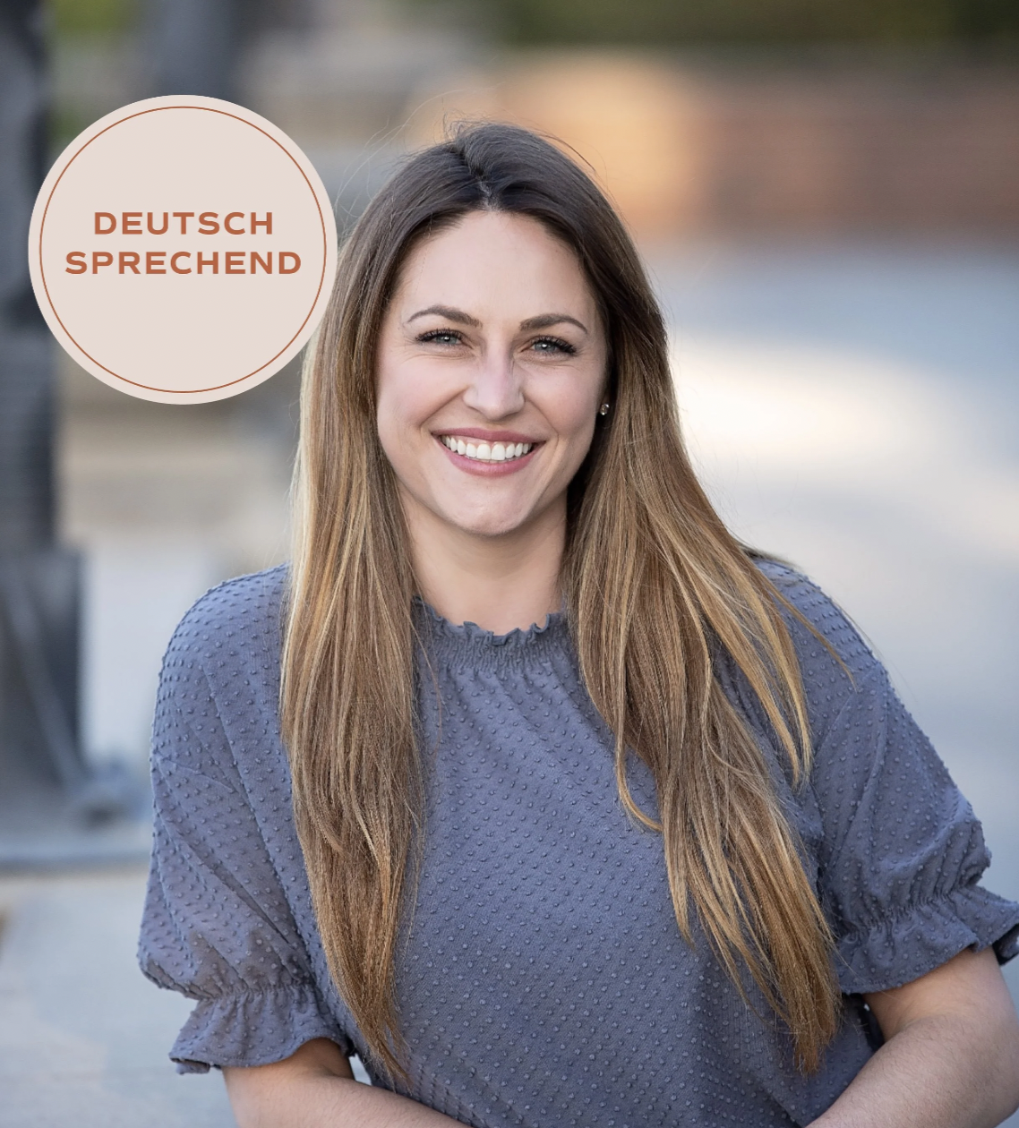 A smiling woman with long brown hair wearing a blue top, outdoors by a body of water. A circular label on the image says "Deutschsprechend" indicating she speaks German.