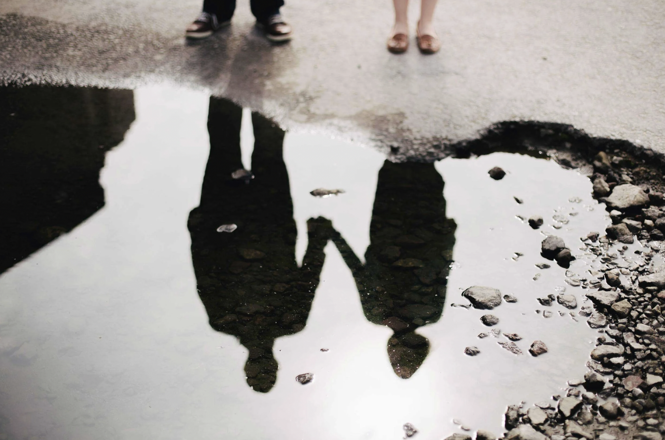 Reflection of a couple holding hands in a puddle on a gravel path, with the top of their bodies visible above.