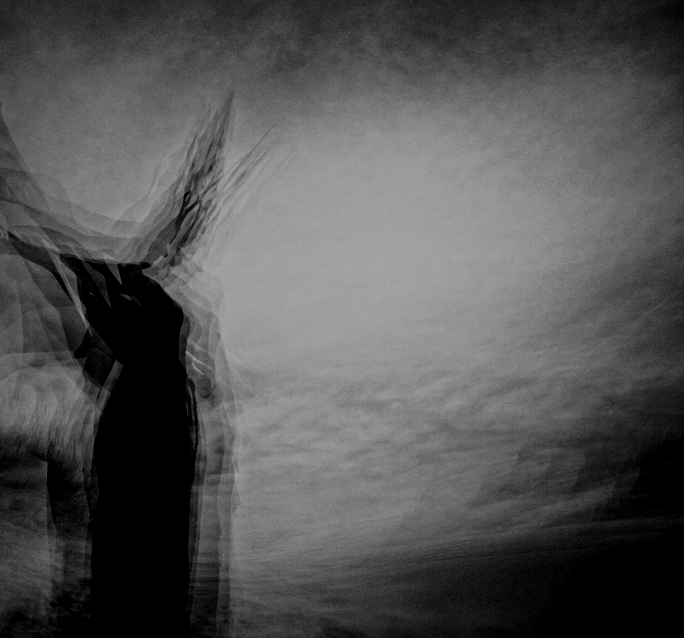 A person with long hair standing on a beach, facing the ocean, captured with a motion blur effect in black and white.